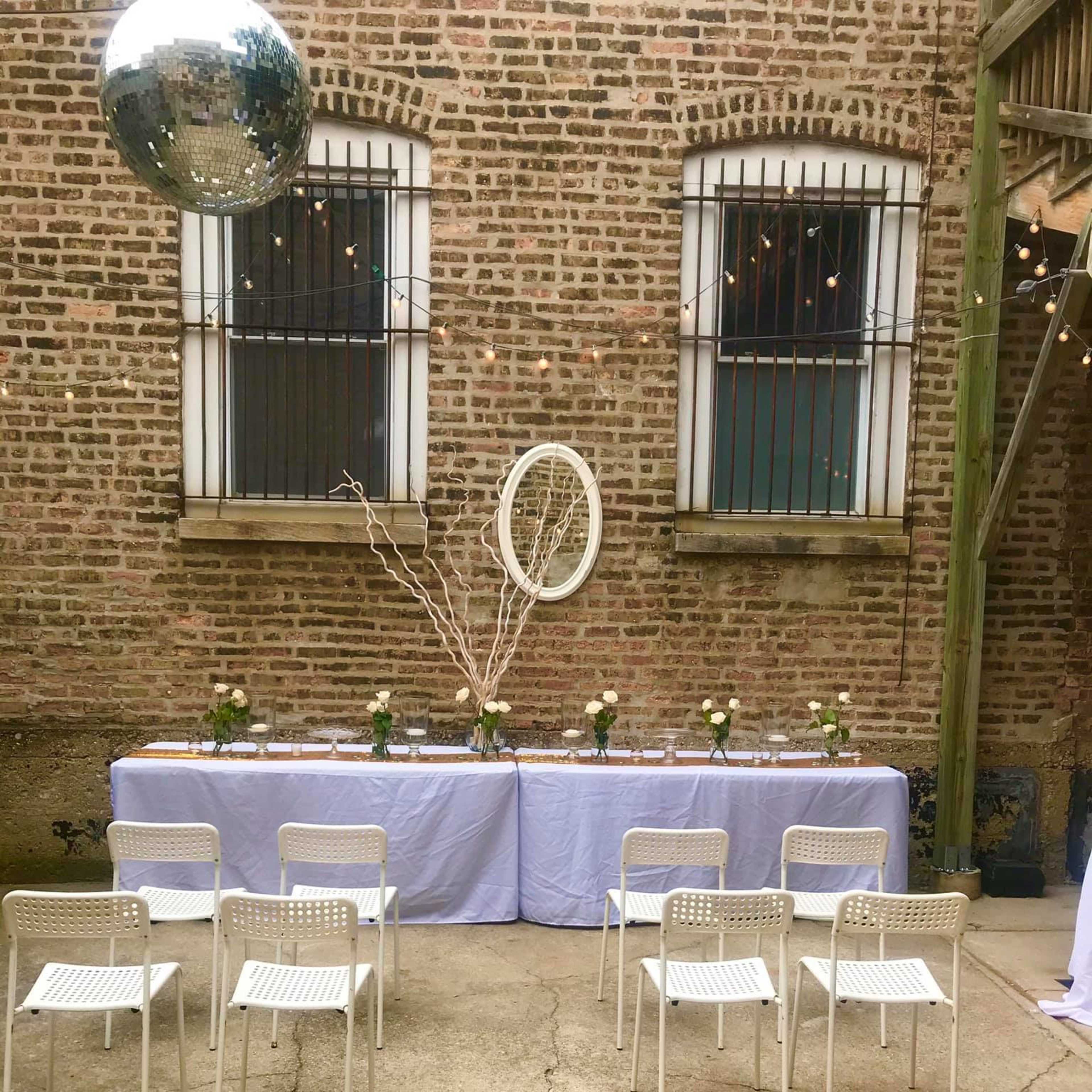 A courtyard setting with a long table draped in white linen, surrounded by white chairs, and a disco ball hanging above.