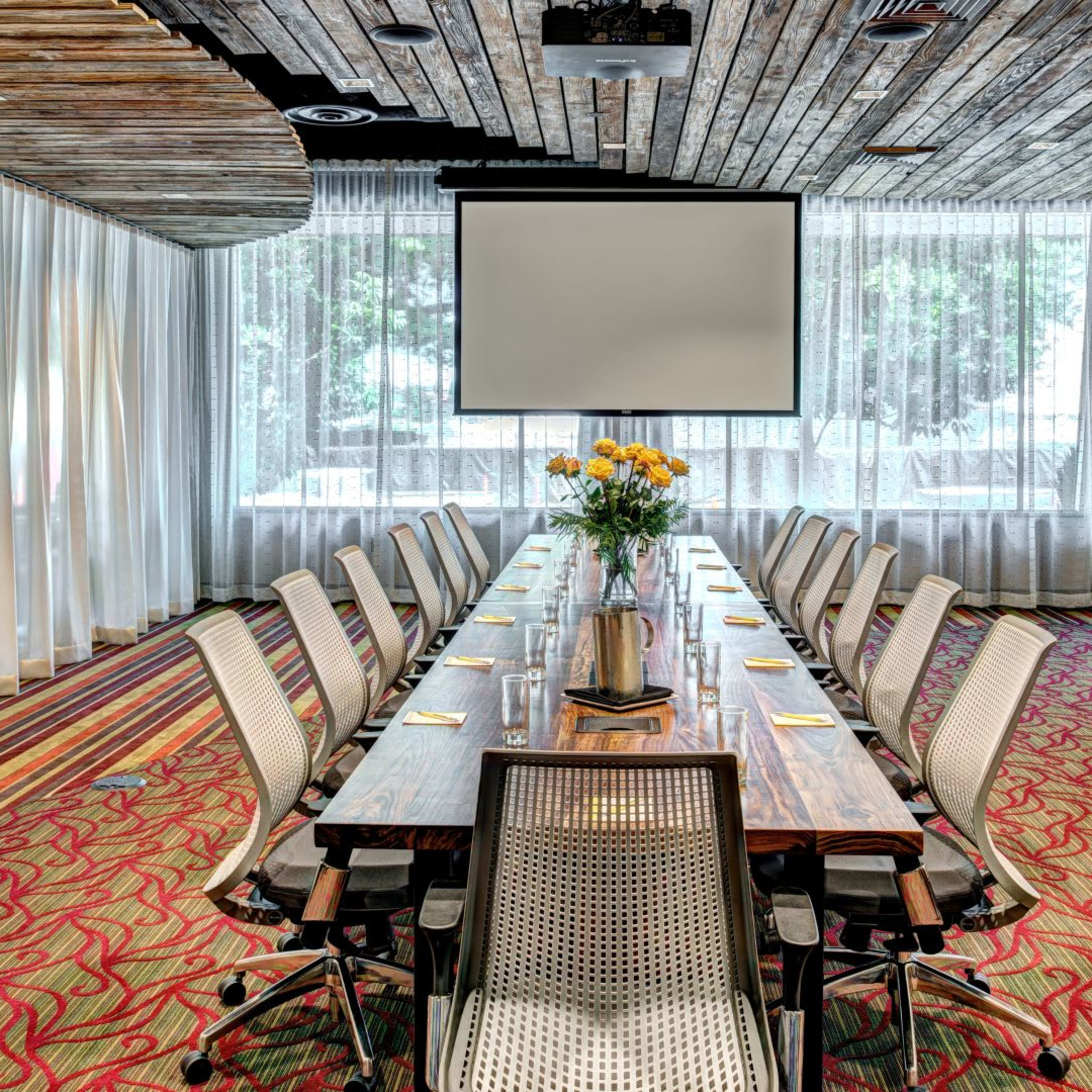 A long wooden conference table is set with water and notebooks, surrounded by ergonomic chairs, in a bright meeting room with large windows and sheer curtains.