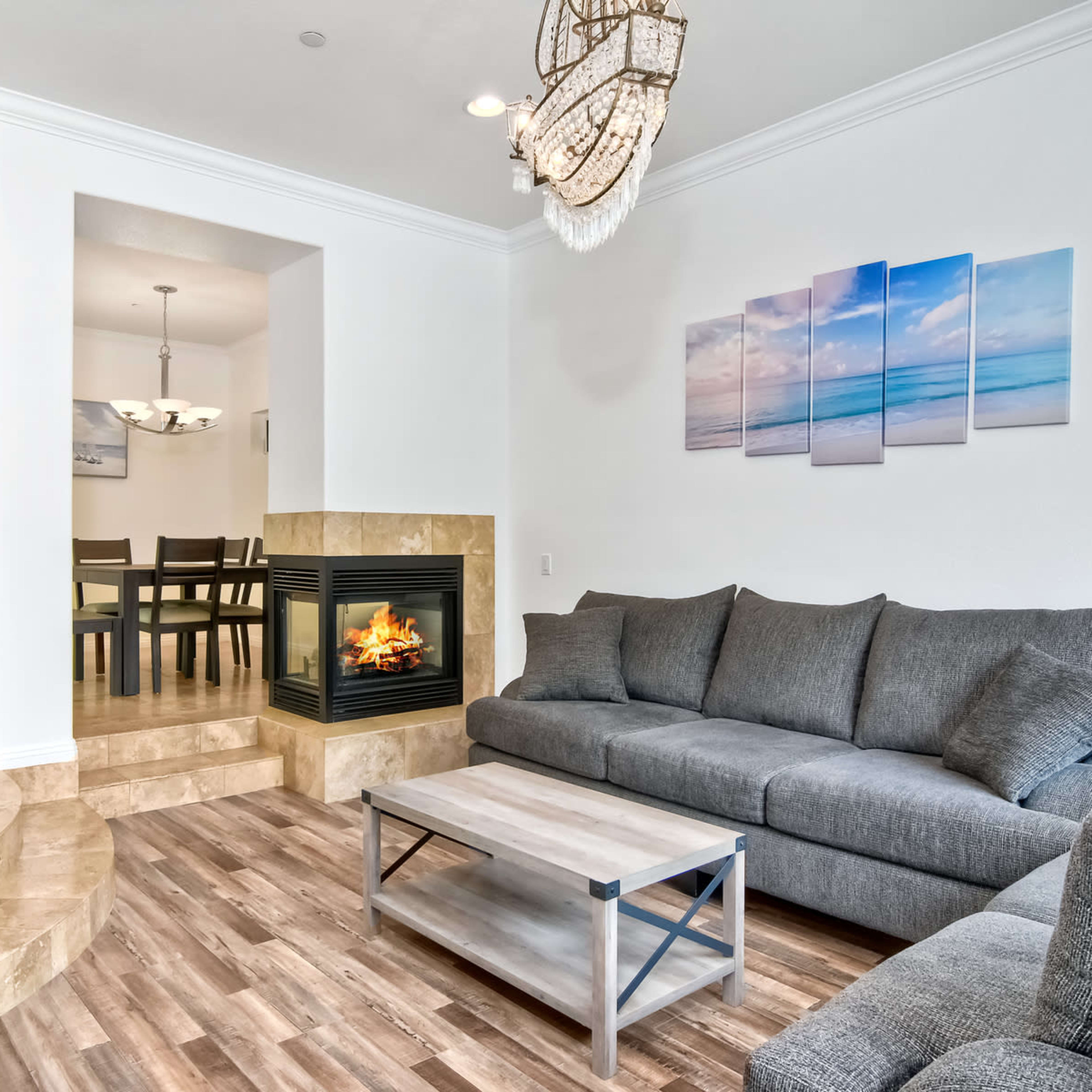 A modern living room with a gray sectional sofa, a coffee table, and a fireplace set into a stone platform, leading to a dining area with a chandelier.