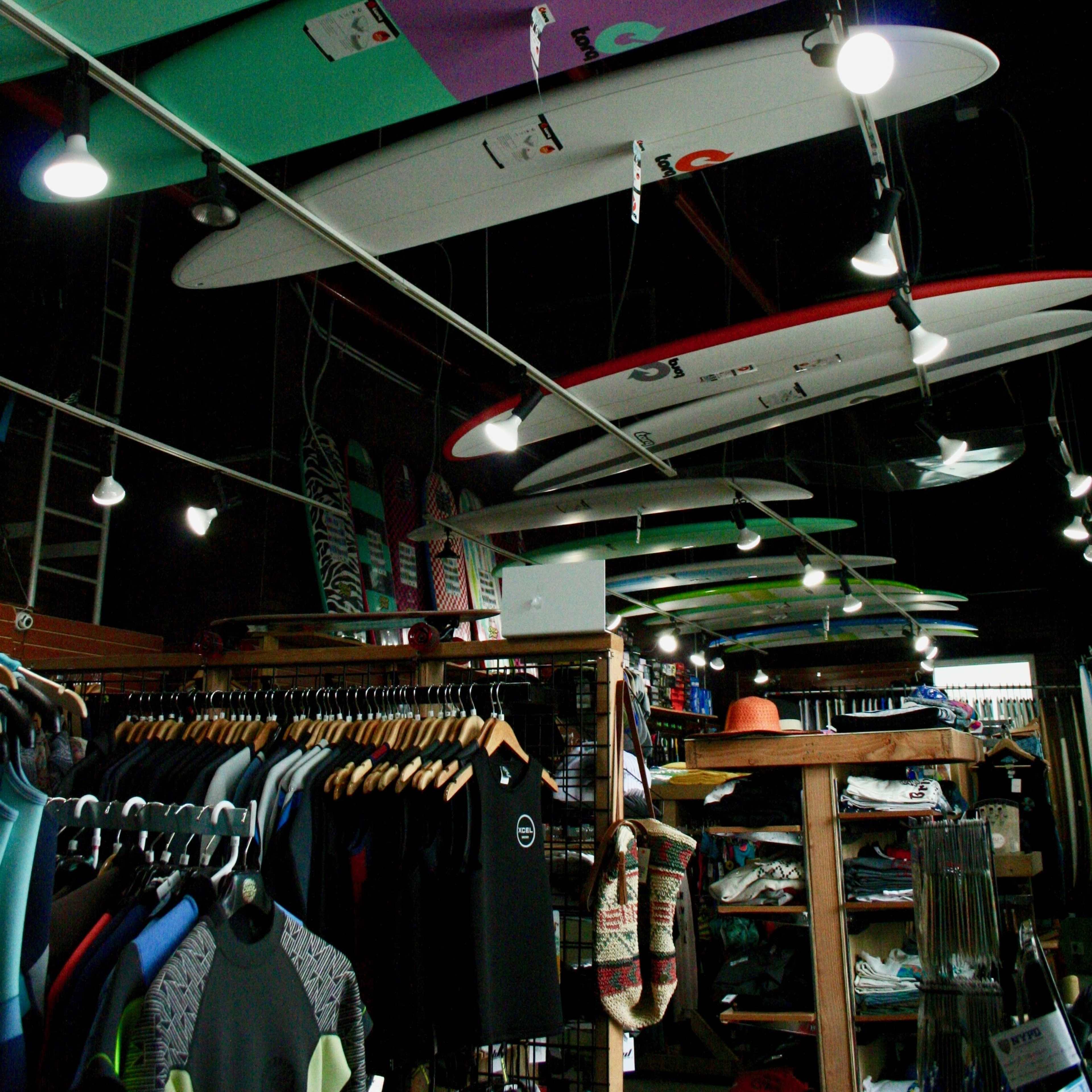 A surf shop displays colorful surfboards hanging from the ceiling, with racks of wetsuits and accessories arranged below.