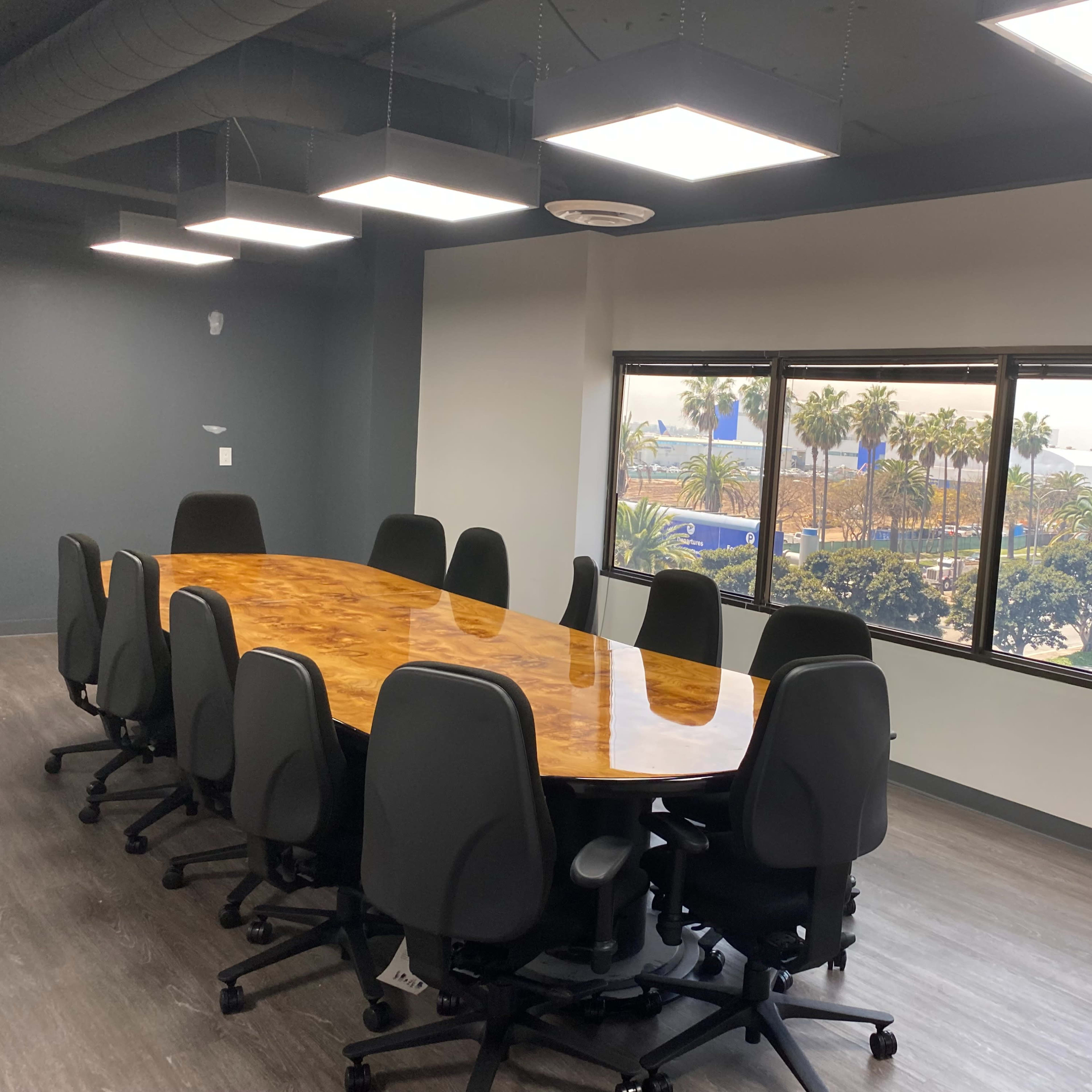 A long wooden conference table surrounded by executive chairs sits in a bright meeting room with large windows overlooking palm trees and a cityscape.