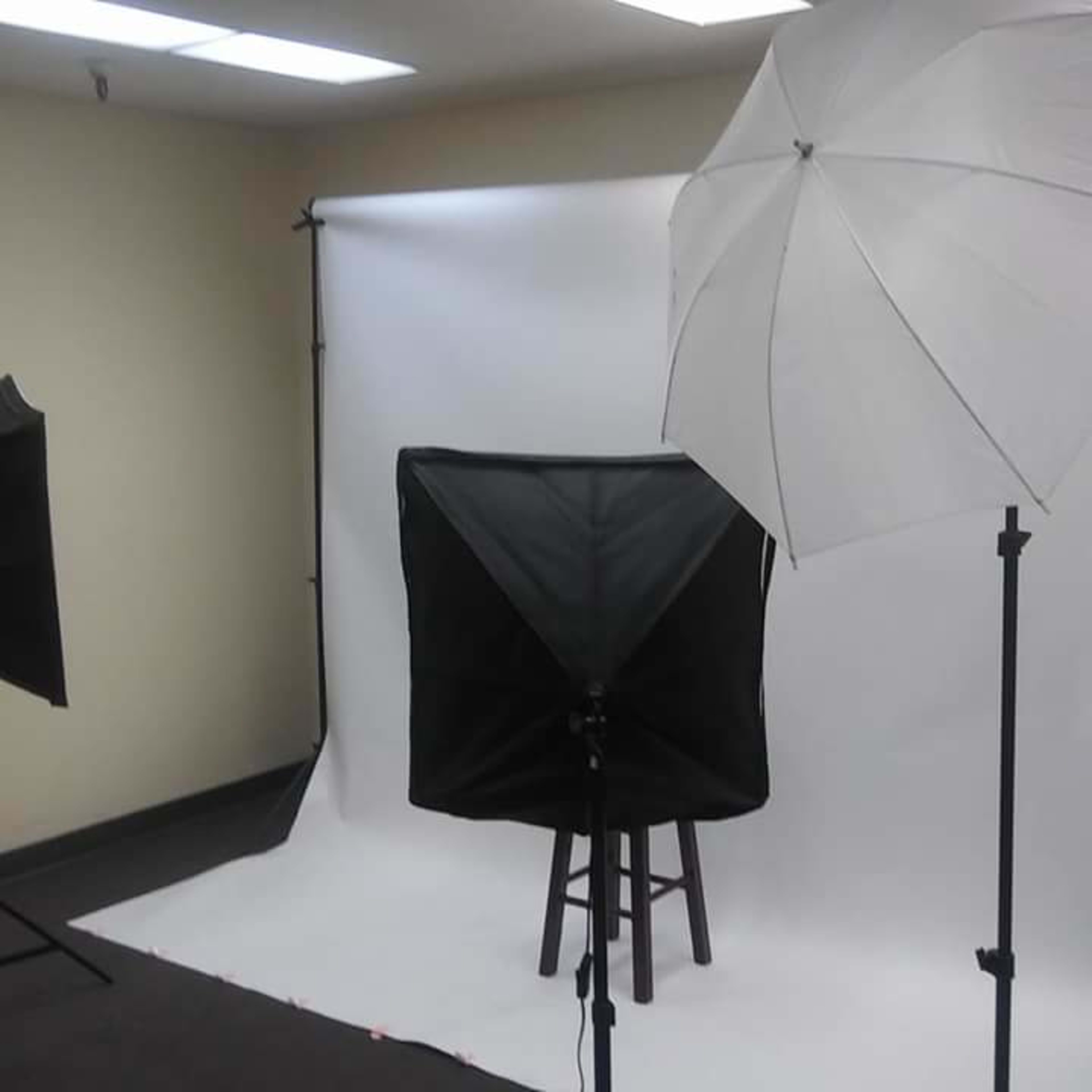A photography setup with two light umbrellas, a black softbox, and a stool in front of a white backdrop.