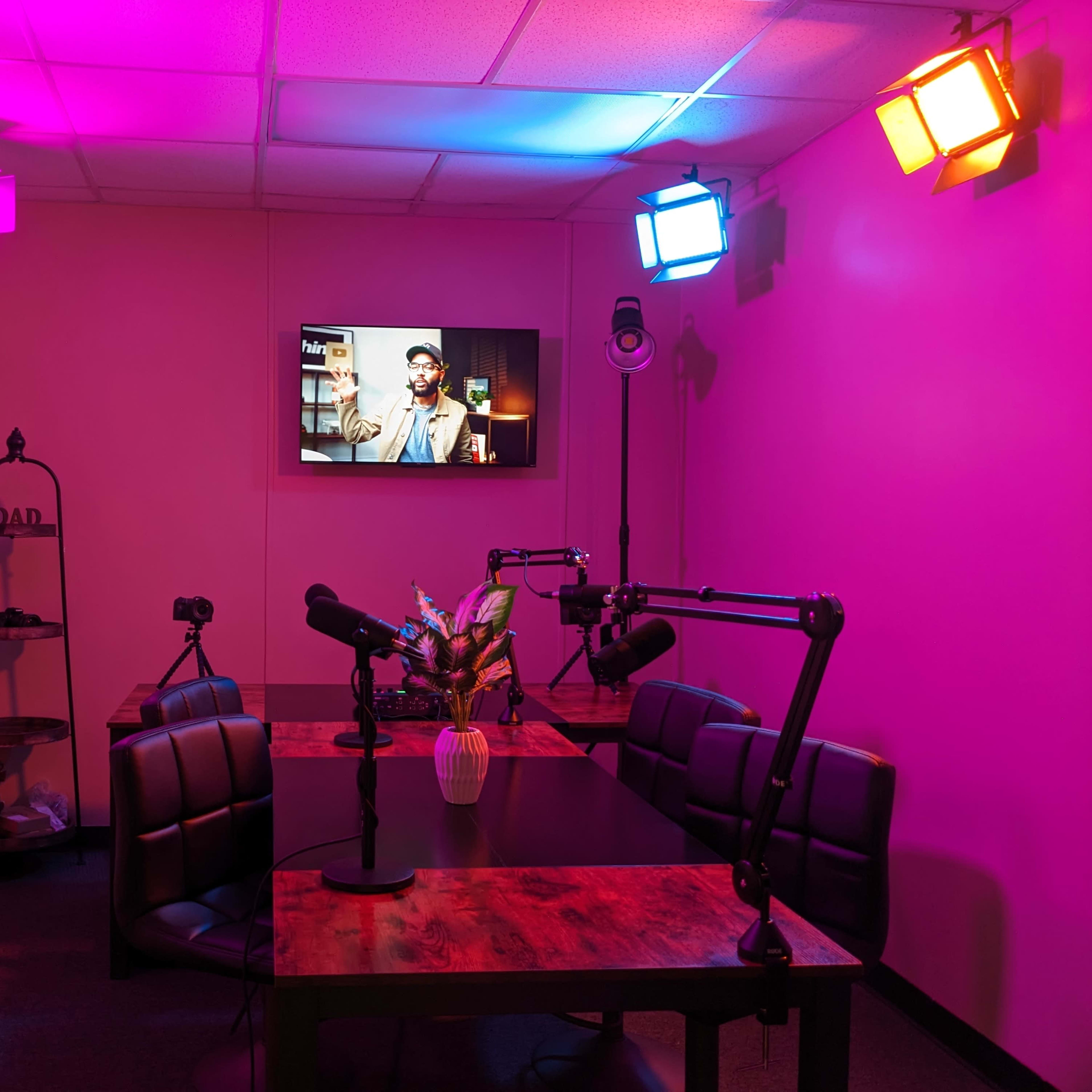 A small recording studio with a table, chairs, and cameras, illuminated by colorful lights and featuring a television screen on the wall.
