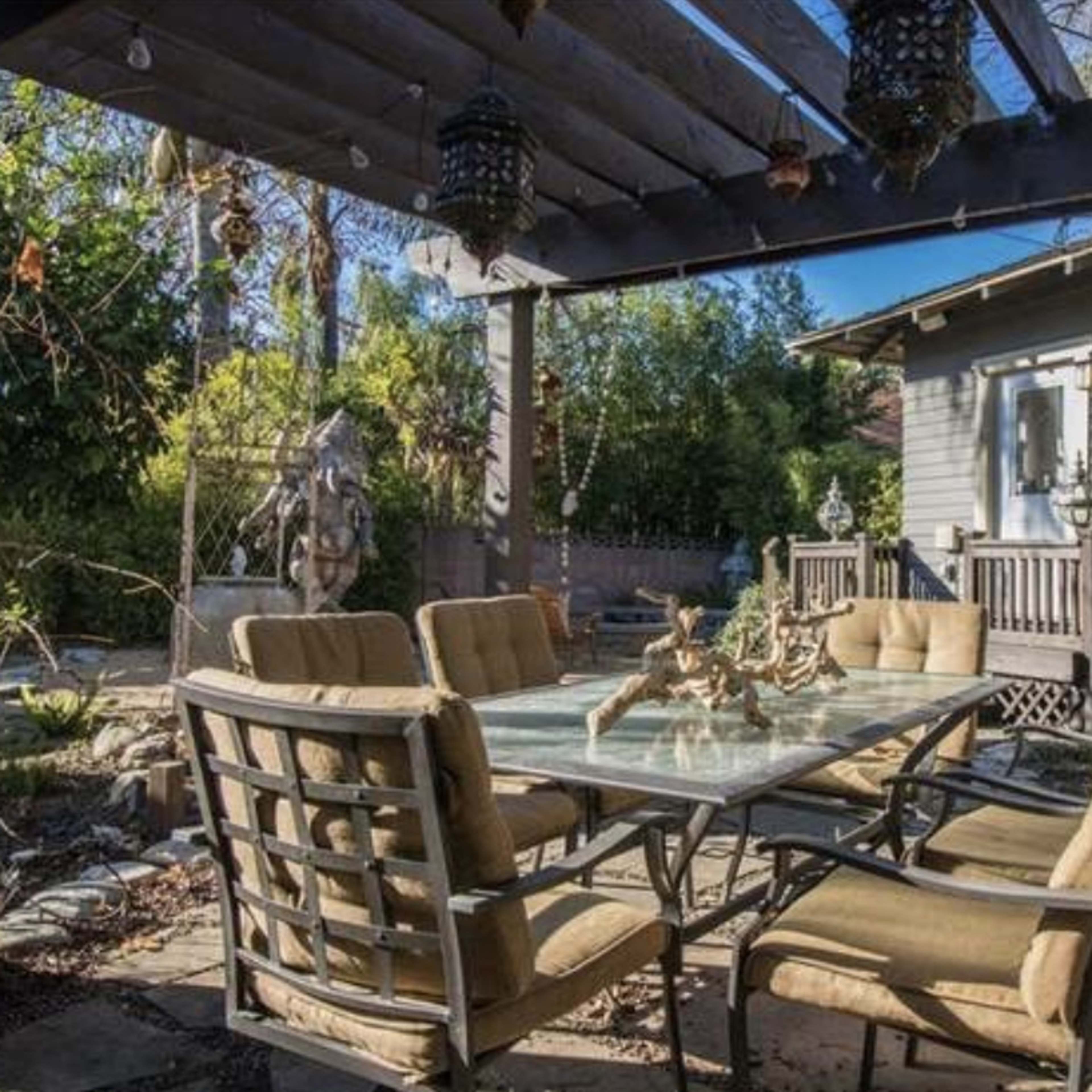 A patio with a glass table and six chairs is shaded by a wooden pergola, surrounded by greenery and a wooden deck.