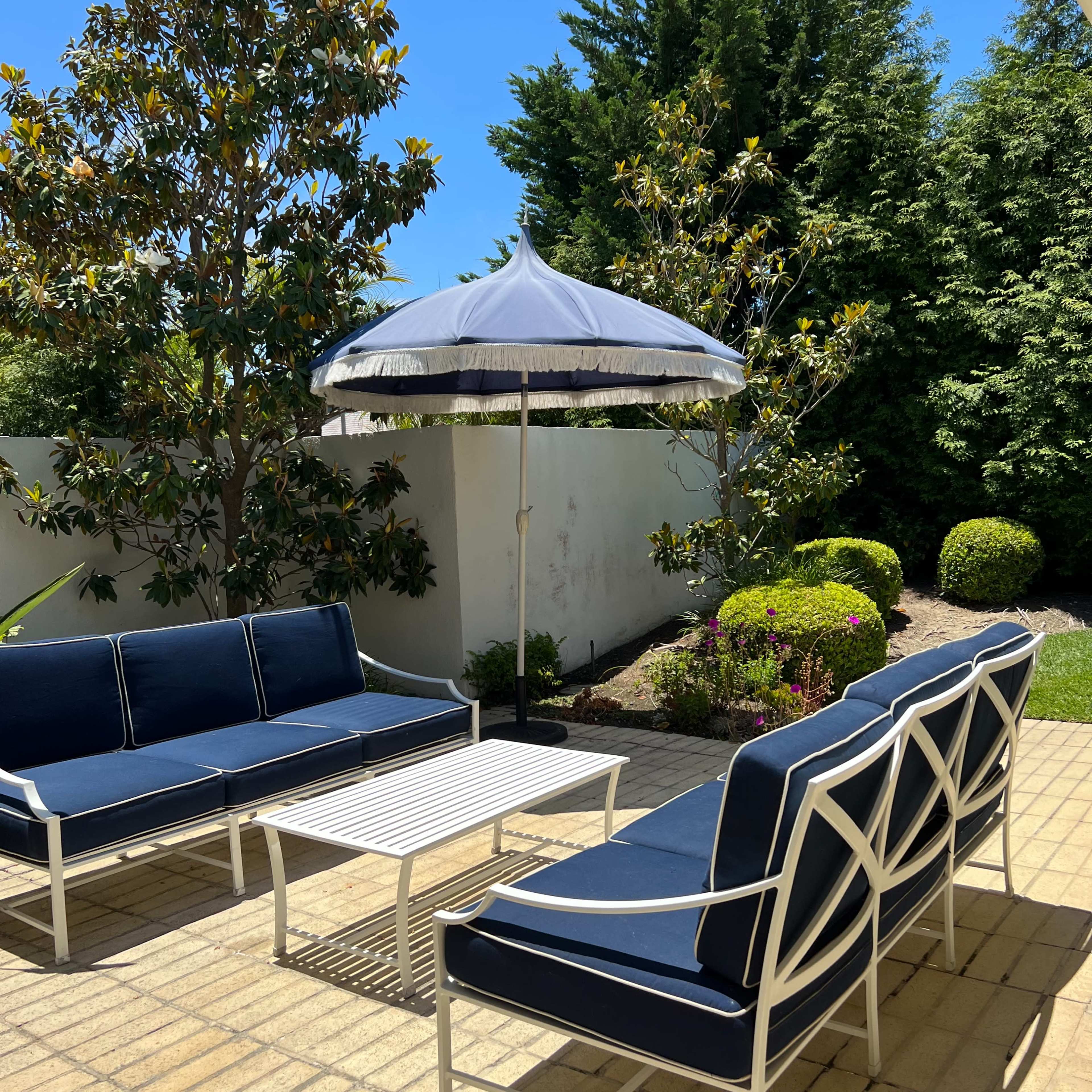 A patio area features navy blue outdoor furniture, including a couch, chairs, and a coffee table, beneath a black and white umbrella, with green shrubs in the background.
