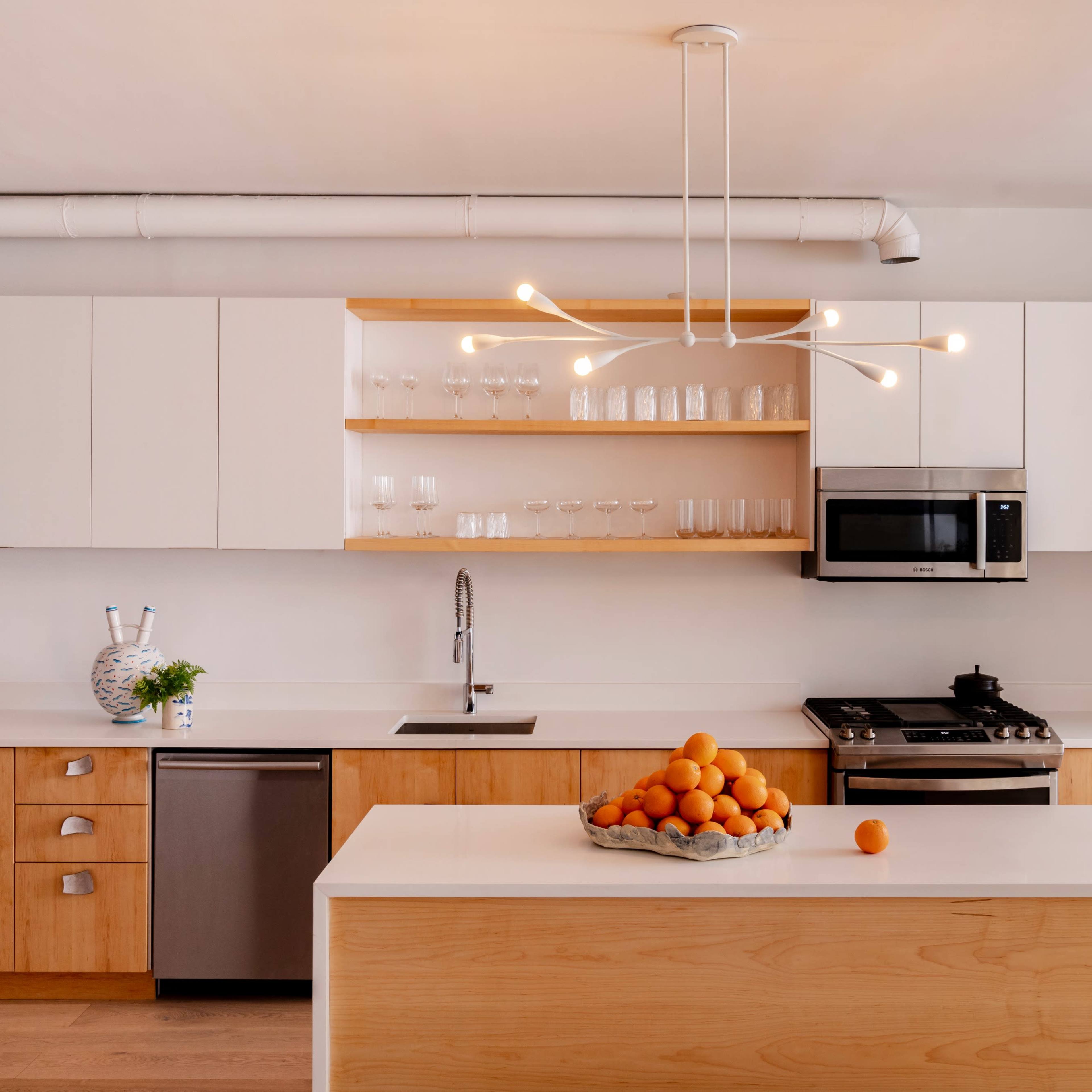 A modern kitchen features light wood cabinetry, a white island with a bowl of oranges, and stainless steel appliances.
