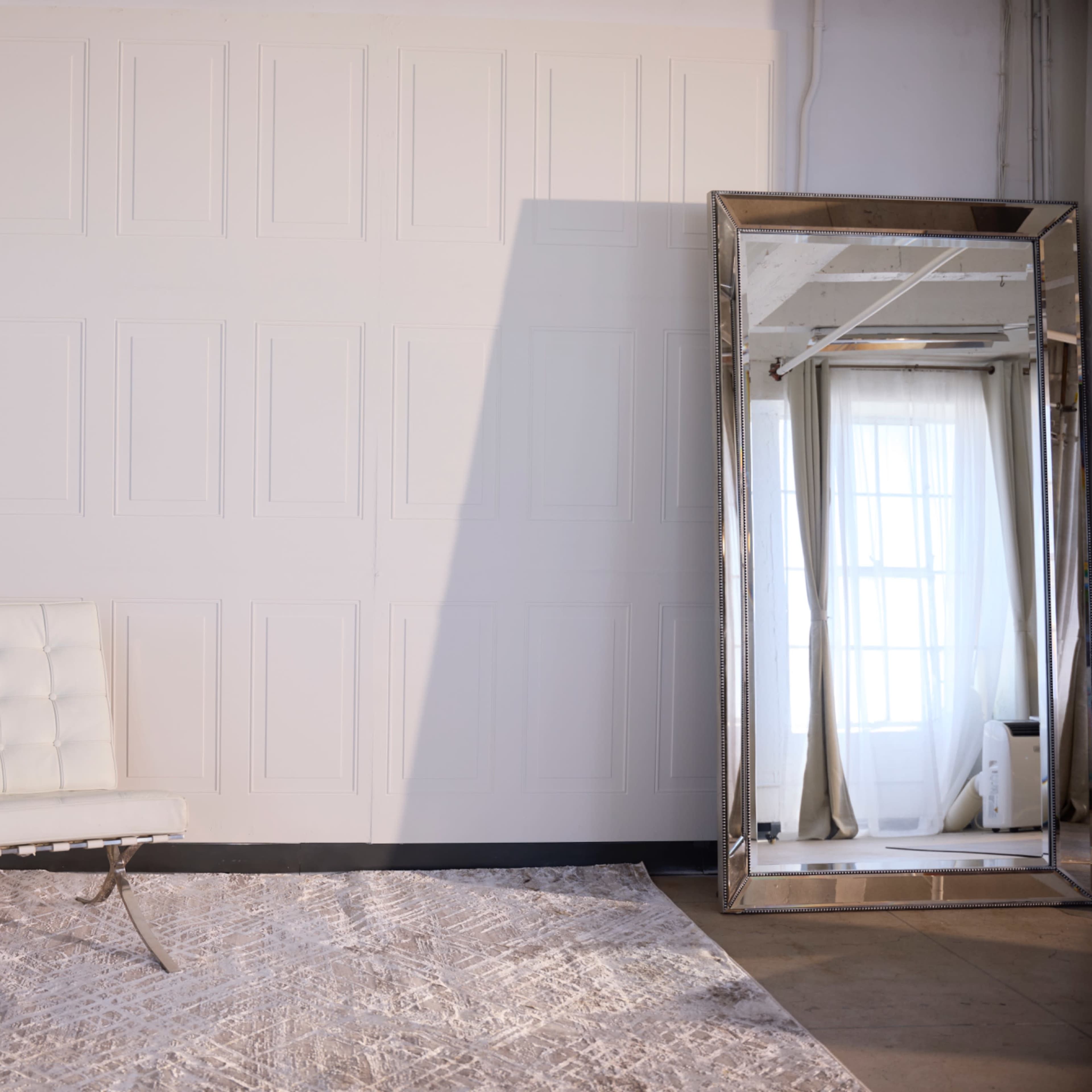 A white tufted chair is placed on a patterned rug next to a large mirror reflecting a window.