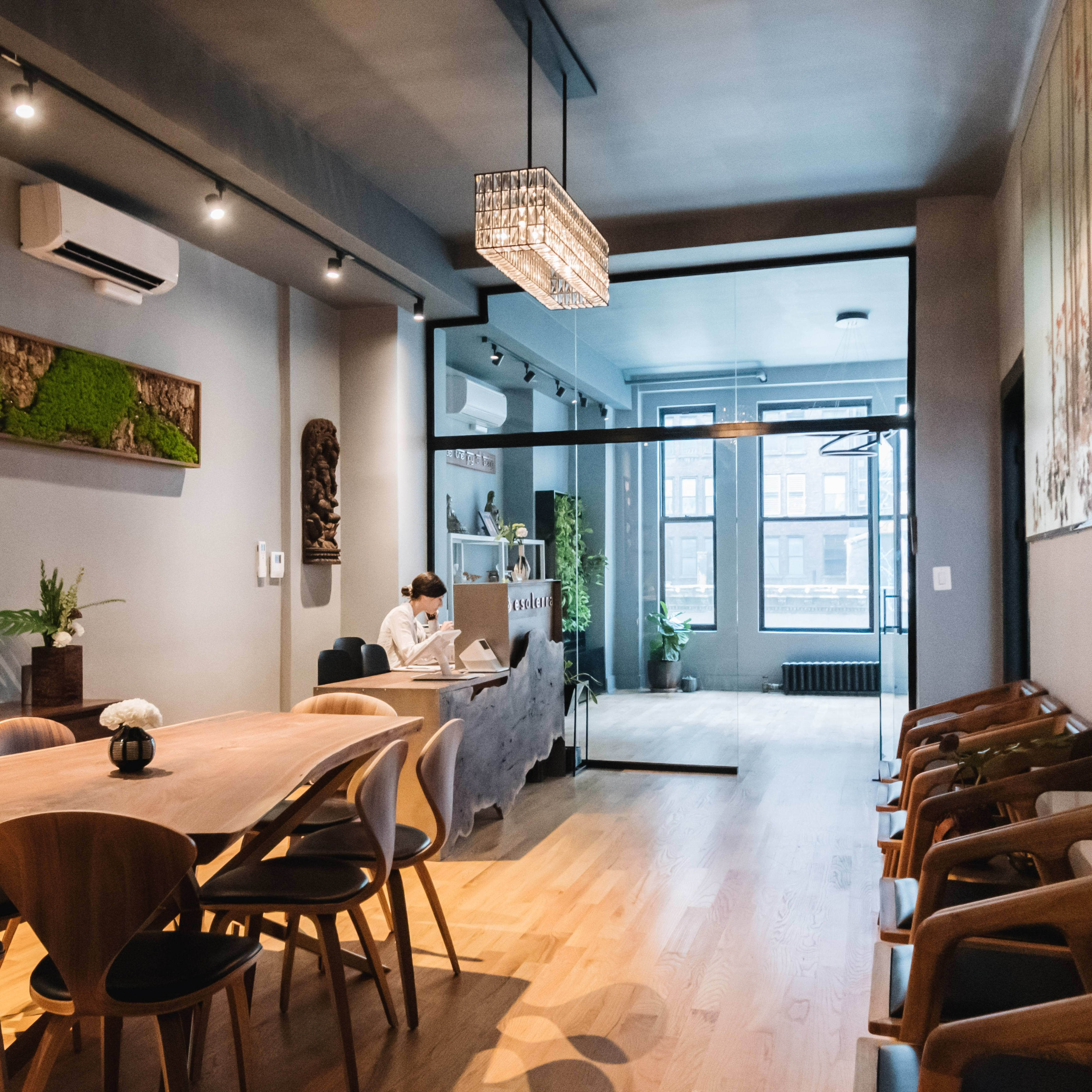 A spacious, modern interior features a long wooden table with chairs, a person working on a laptop, and decorative elements on the walls.