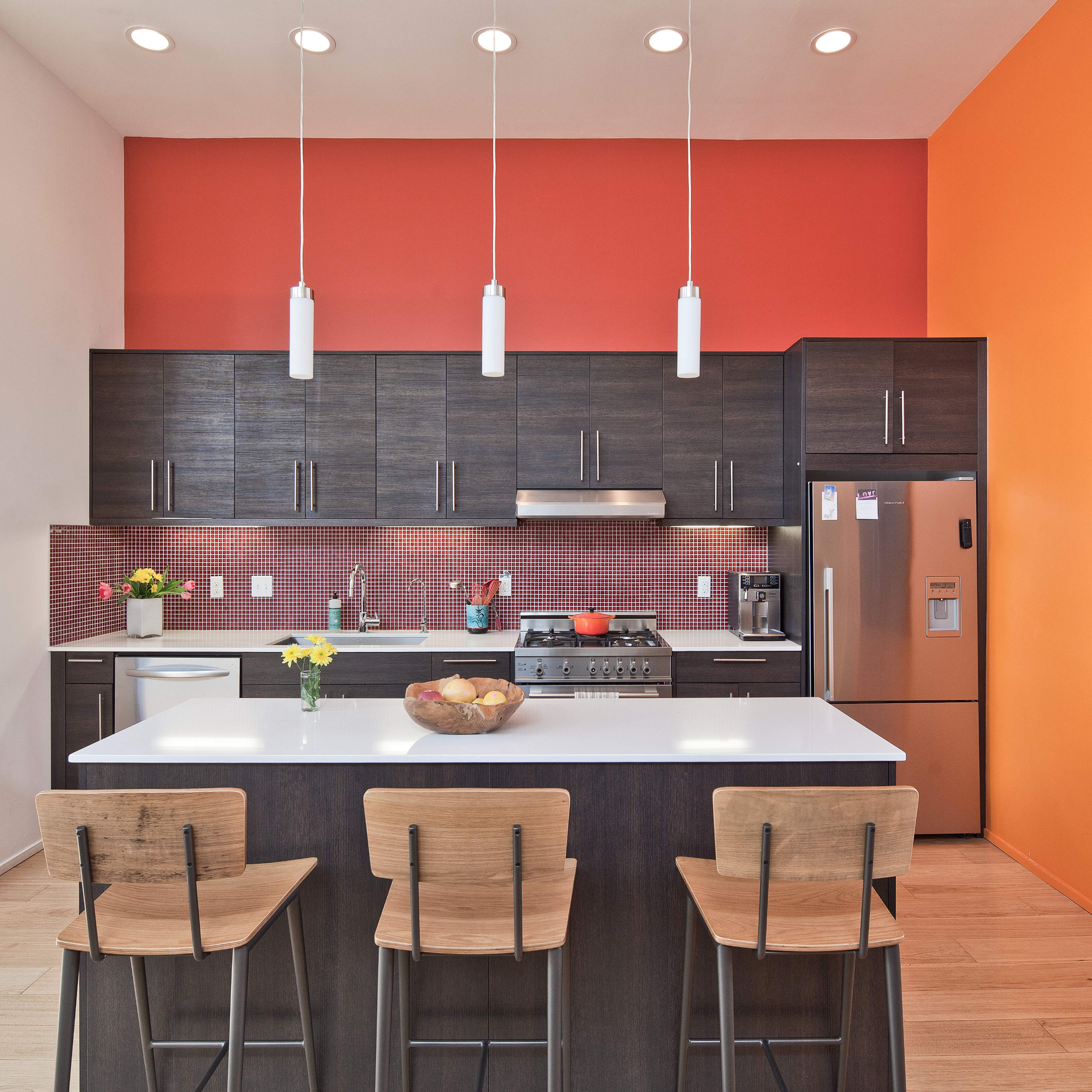 A modern kitchen features dark cabinetry, stainless steel appliances, and a bright orange accent wall with bar stools at the island.