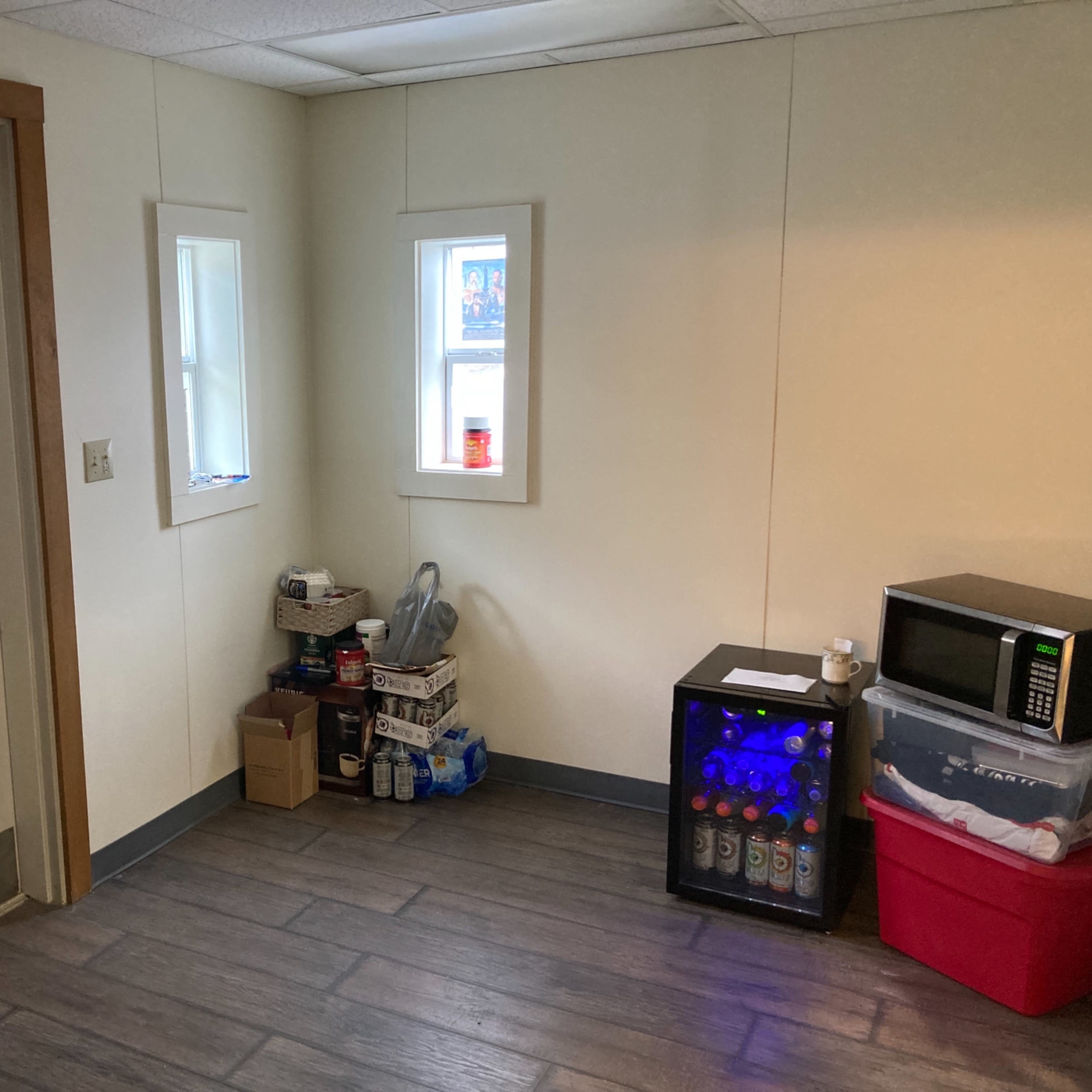 A small corner of a room with a door, two windows, a microwave, a small beverage cooler, and various boxes stored on the floor.