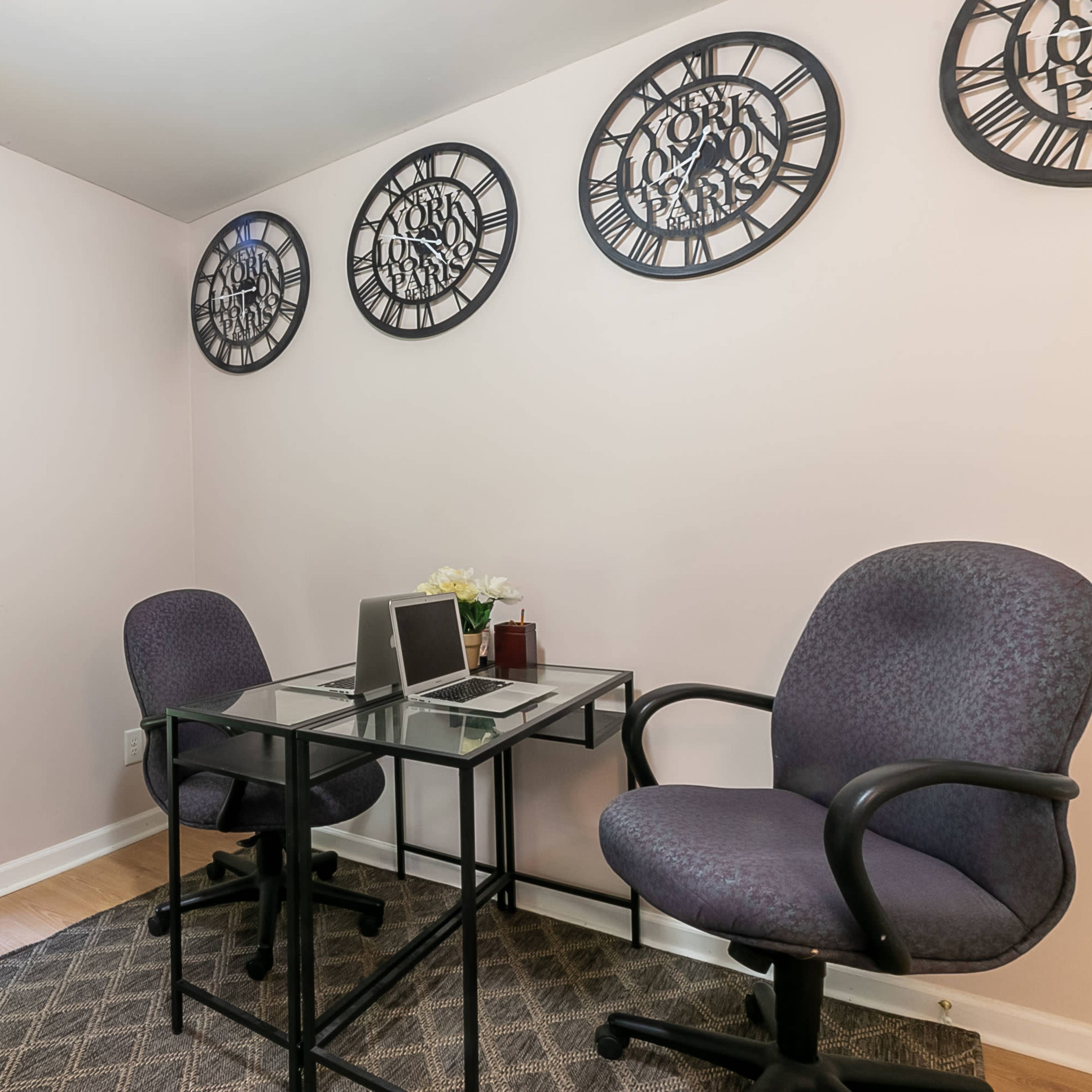 A small office space with two chairs, a glass table, and wall-mounted clocks above it.