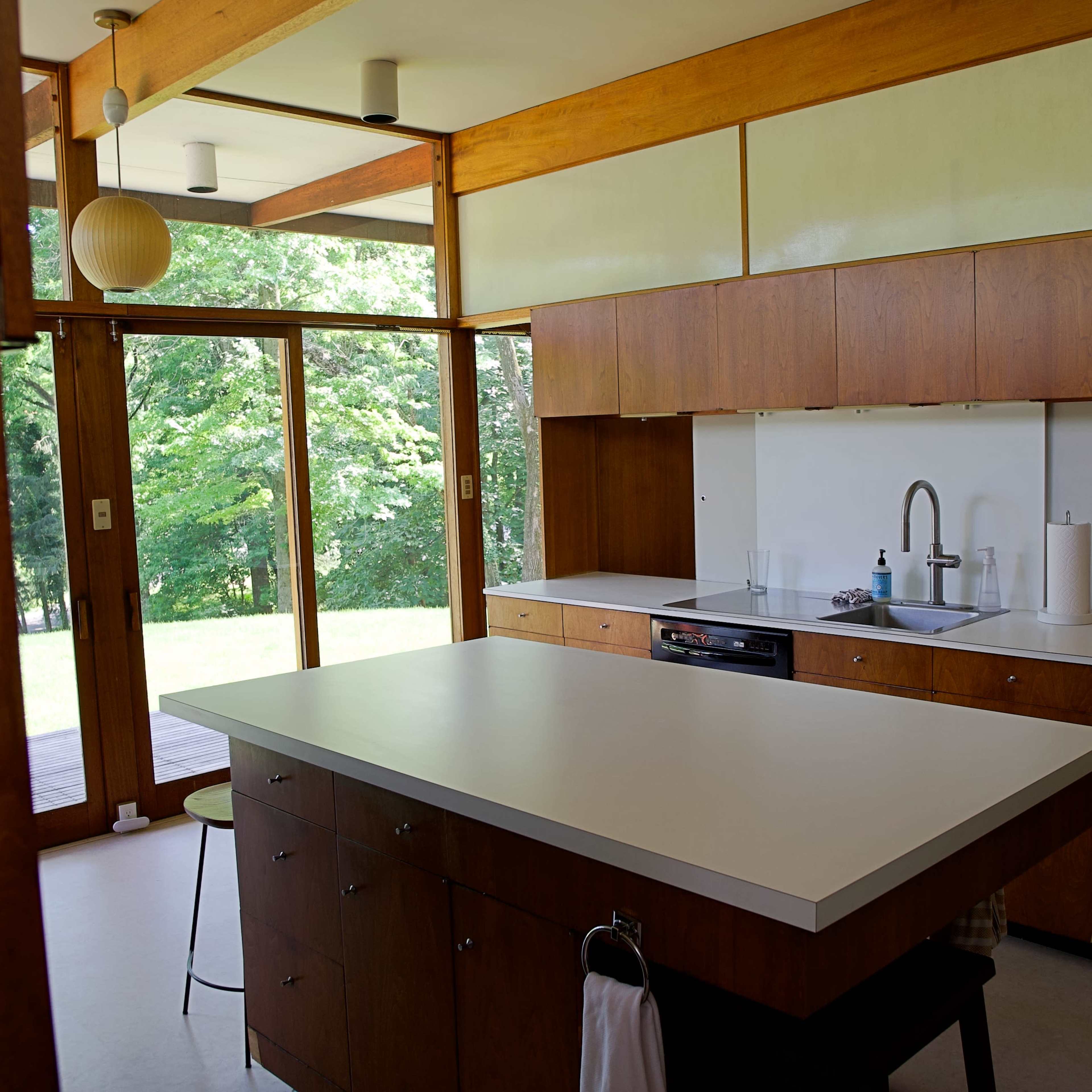 A modern kitchen featuring wooden cabinetry, an island with a white countertop, and large windows that open to a view of a green landscape.