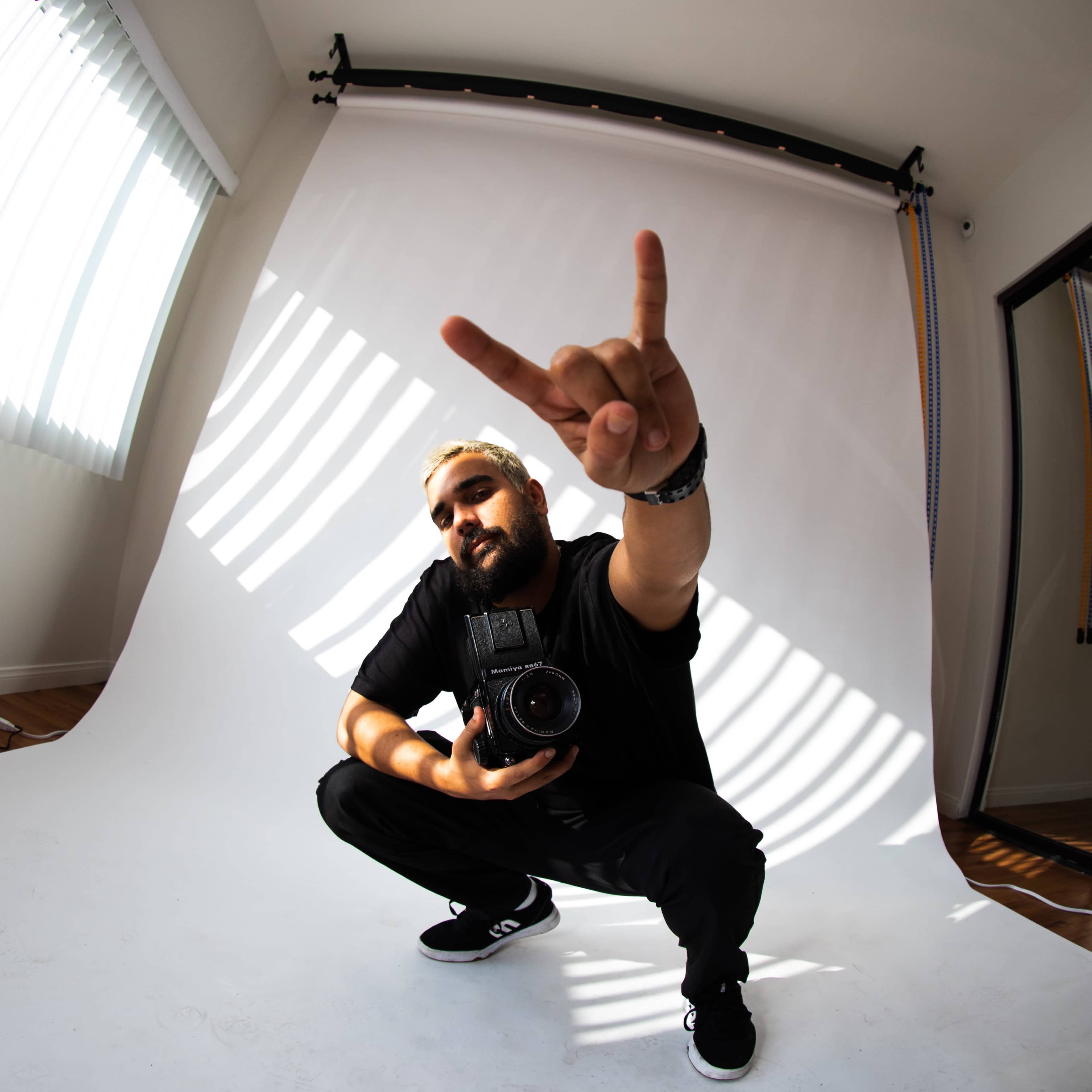 A man in a black shirt and cap squats while holding a camera and making a hand gesture against a white backdrop.