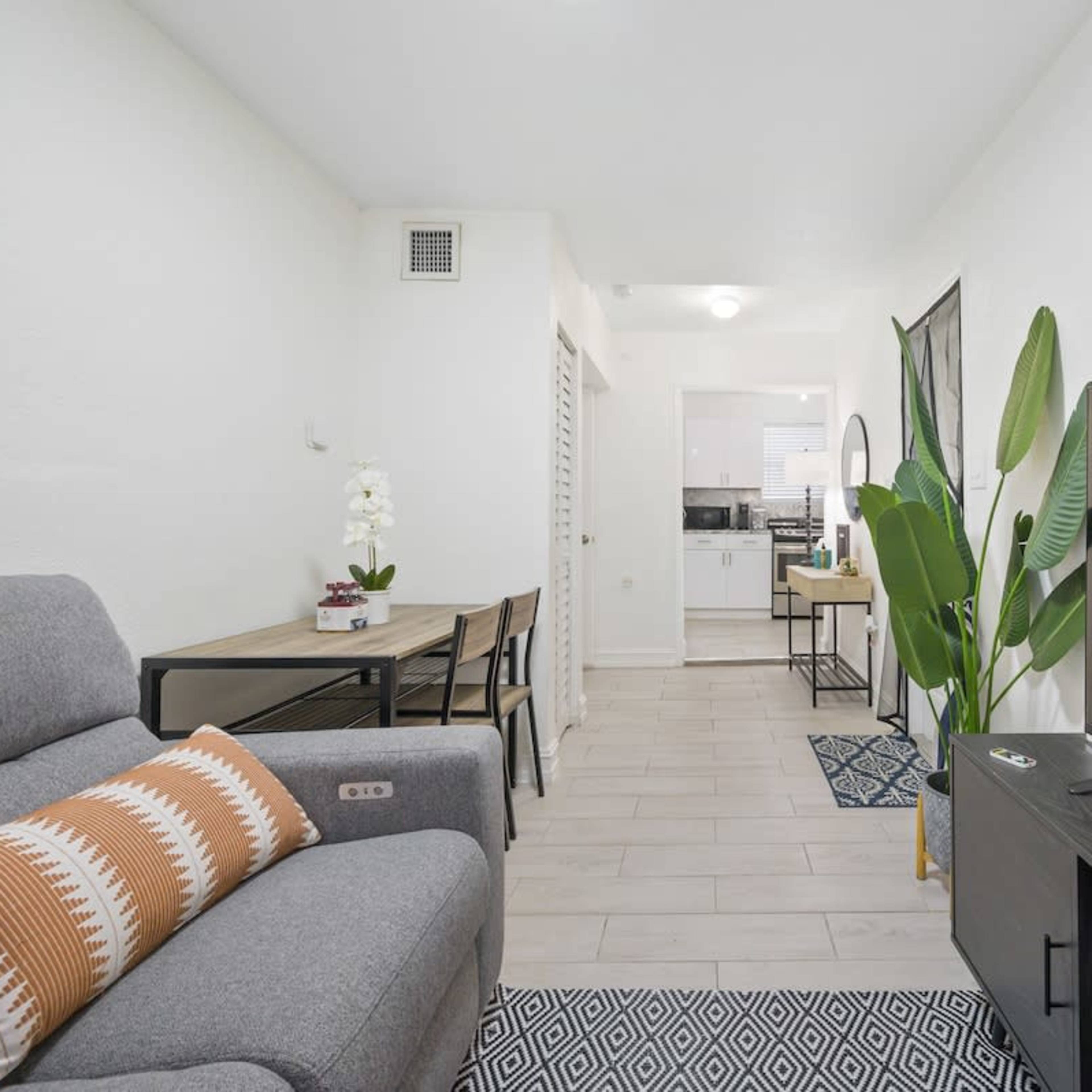 A modern living space with a gray sofa, a dining area, and a television set, featuring large windows and indoor plants.