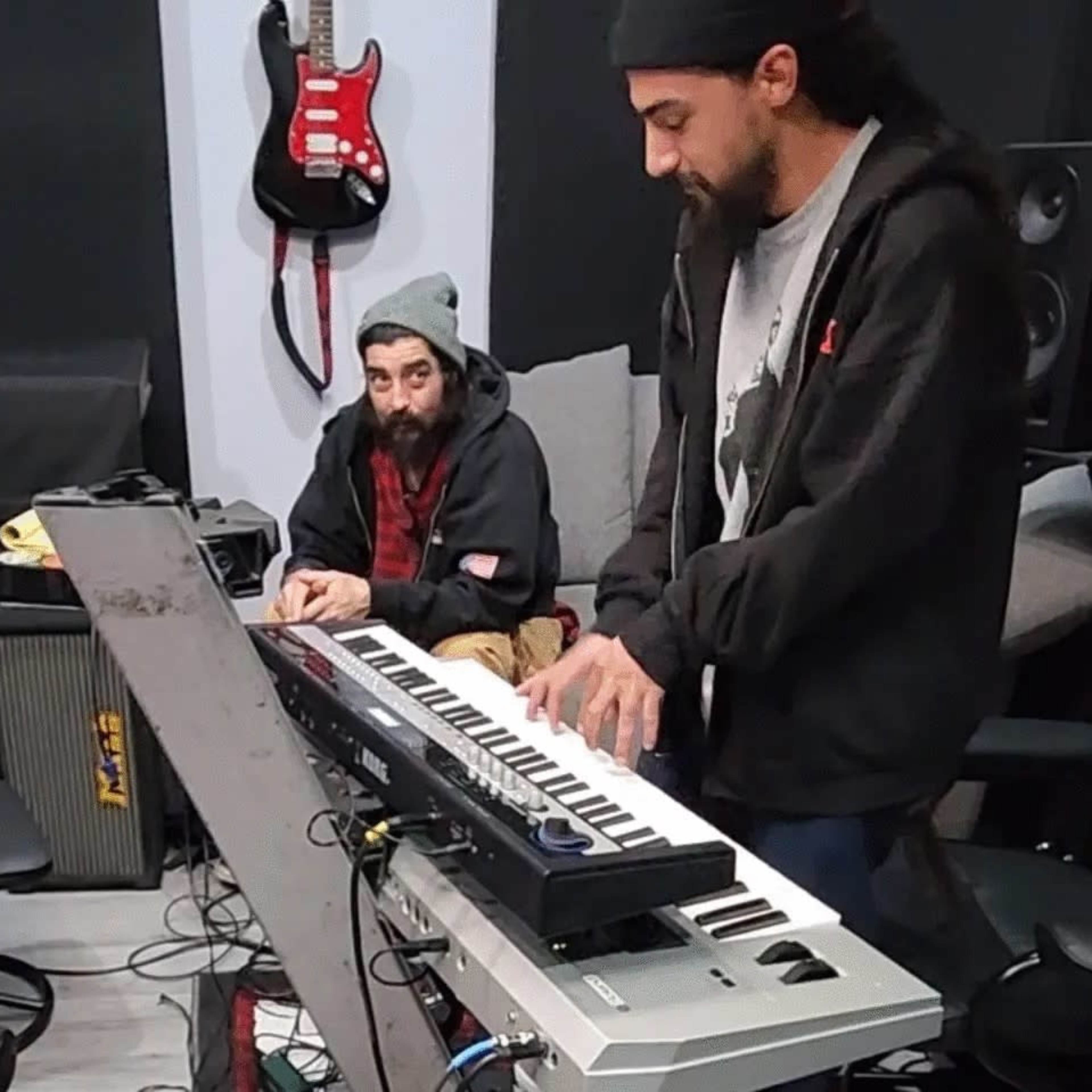 Two men are in a music studio; one is playing a keyboard while the other sits at a table.