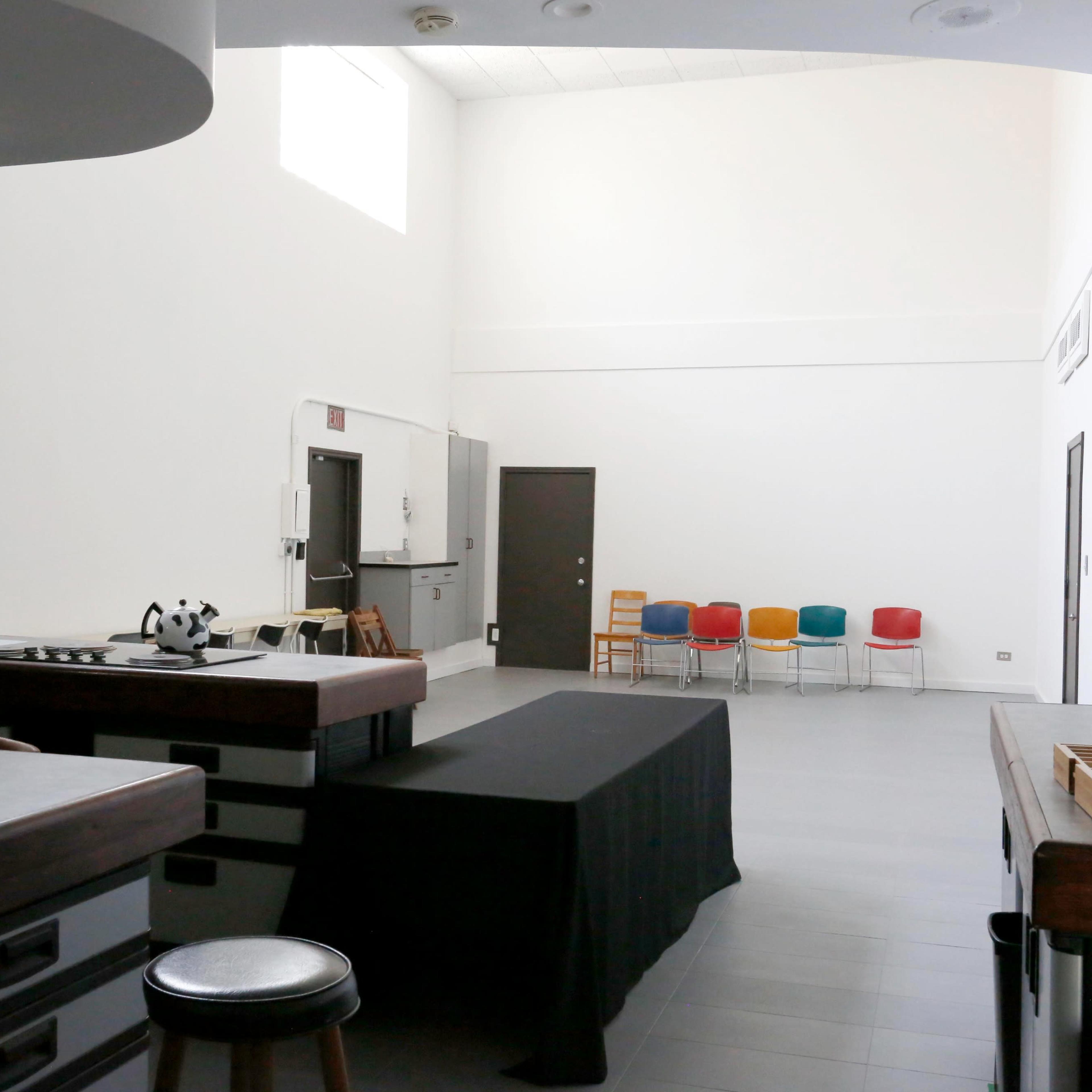 A spacious kitchen area features a black table, a few cooking appliances, and a row of colorful chairs against the wall.