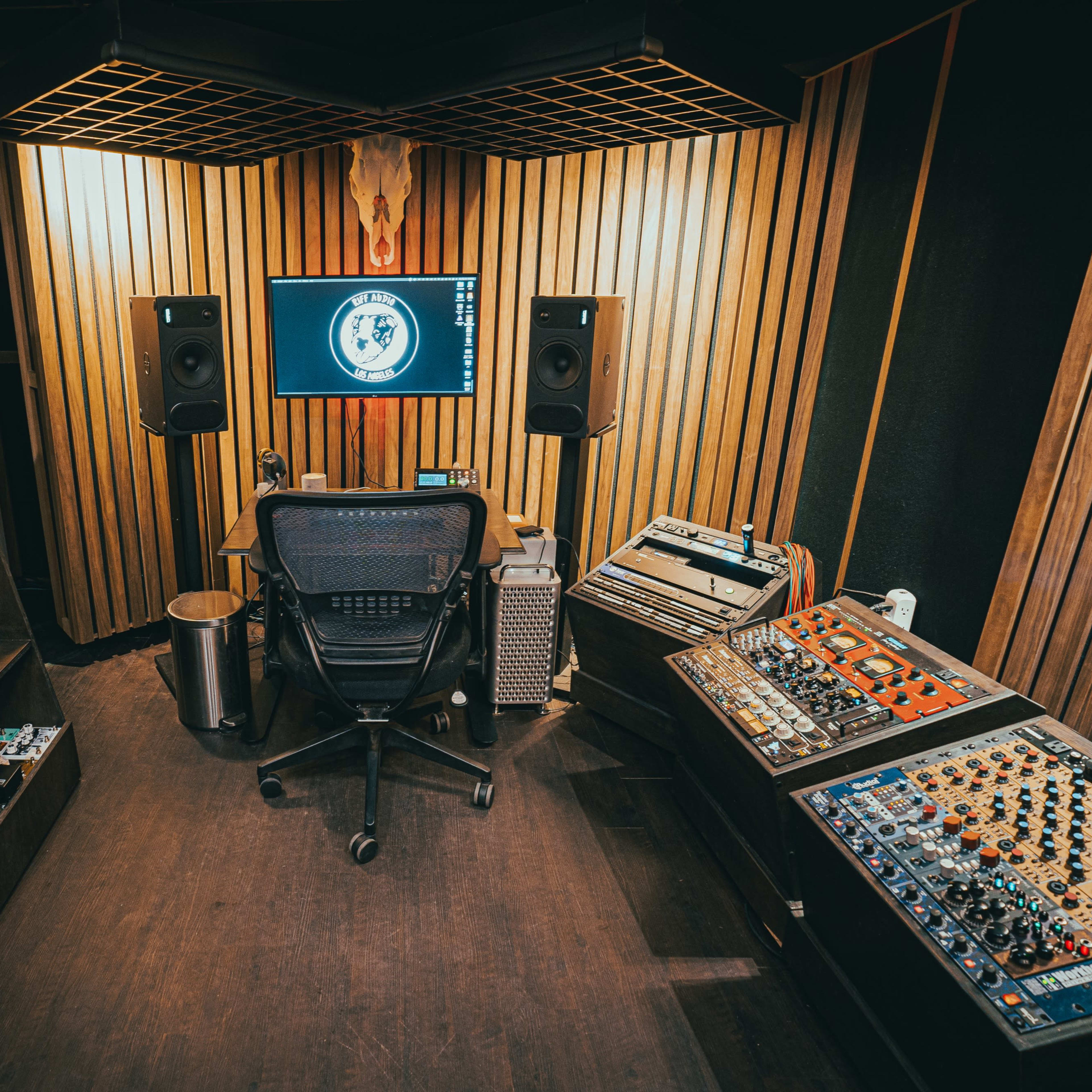 A music production studio features a central chair facing a screen, surrounded by various audio equipment and amplifiers along the walls.