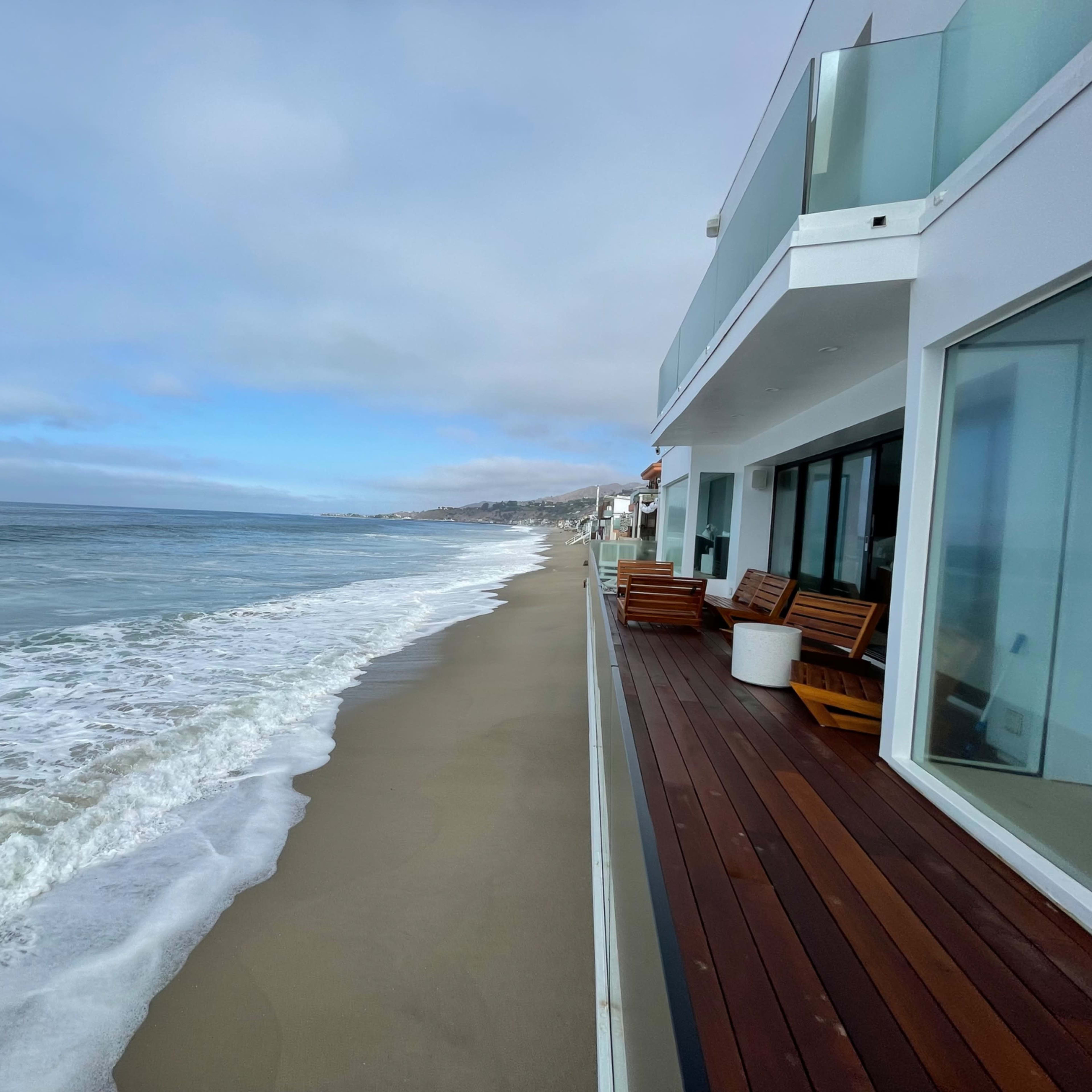 A modern beachfront property overlooks a wide sandy shoreline and gentle waves under a cloudy sky.
