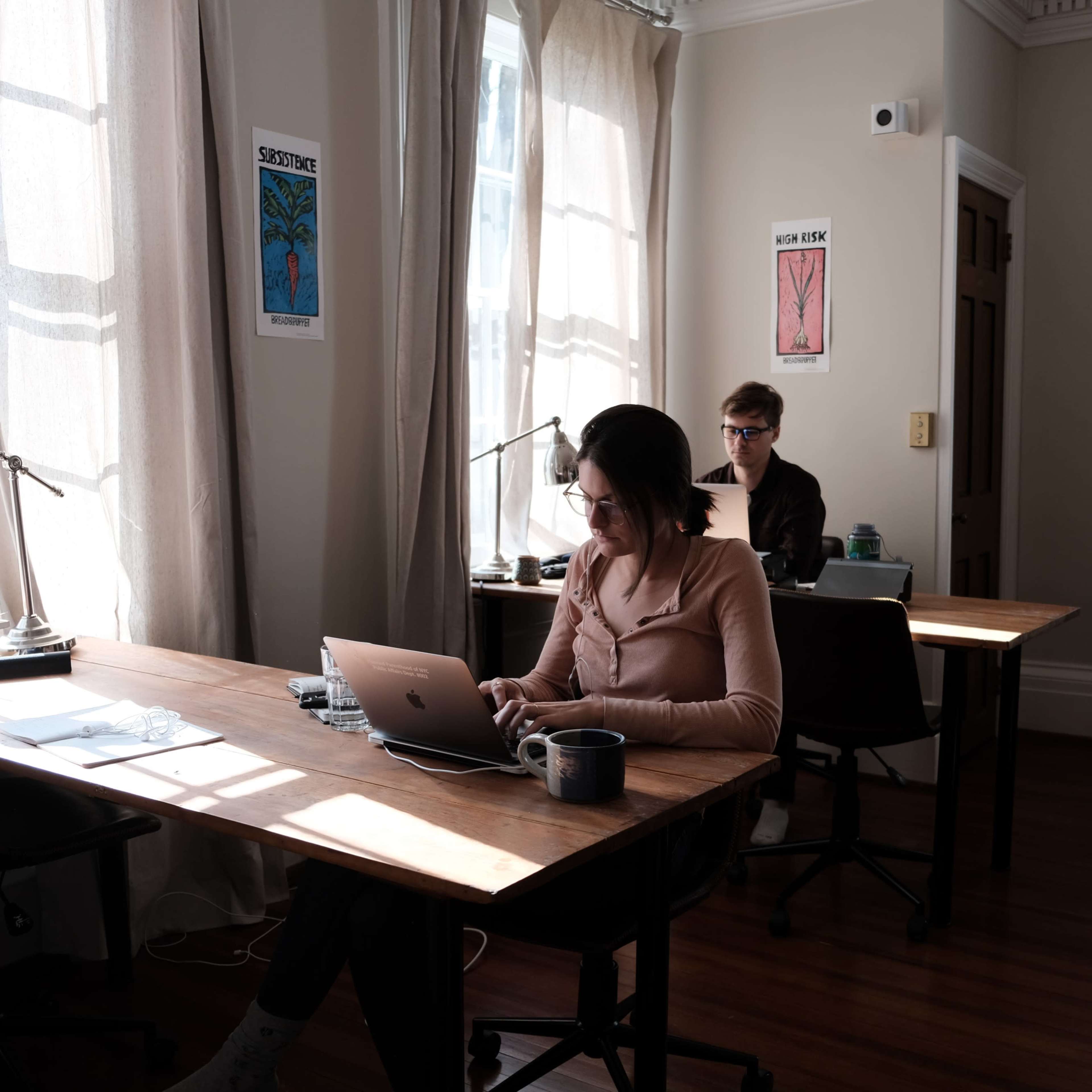 Two people work at separate desks in a well-lit room with large windows and posters on the walls.