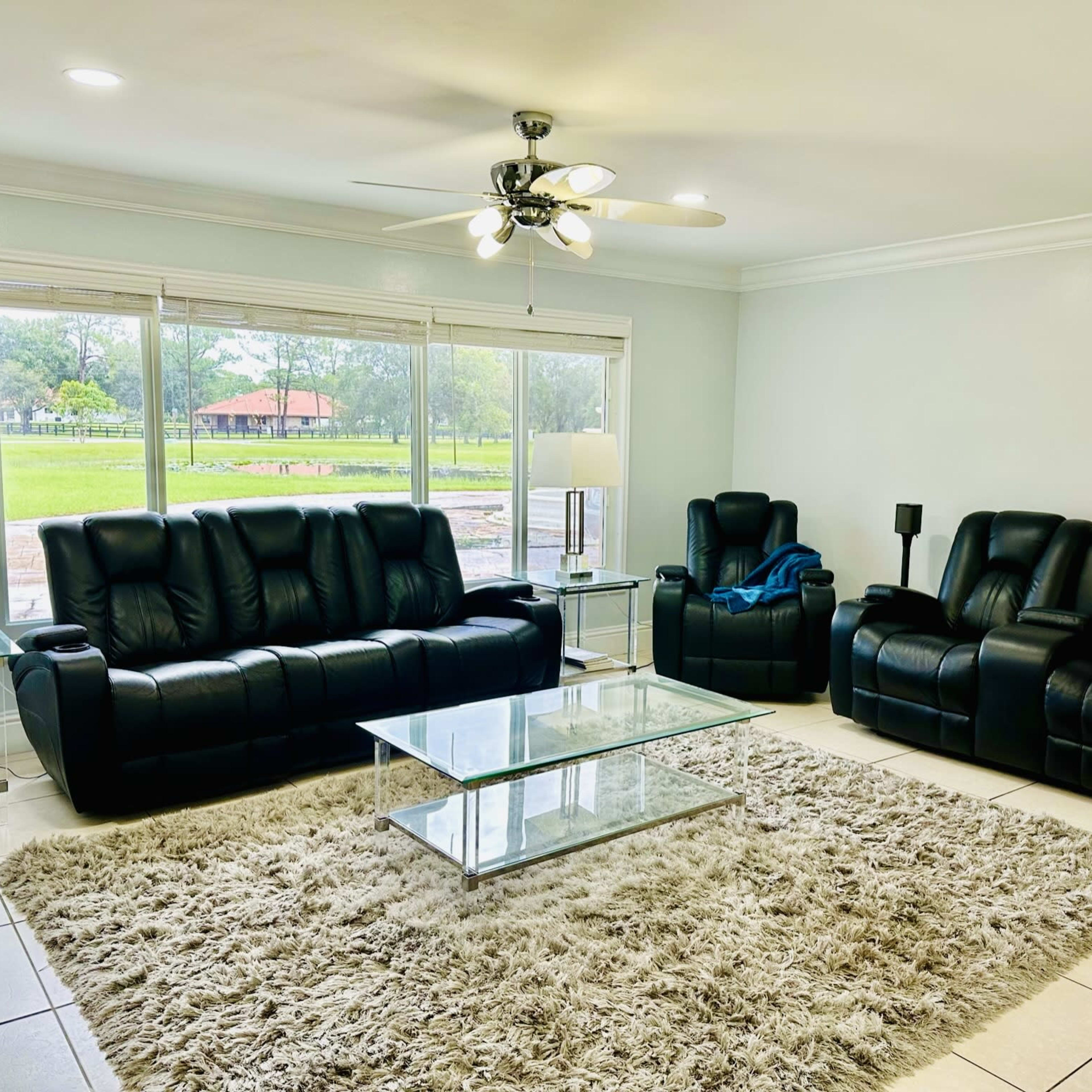 A living room with black leather sofas, a glass coffee table, and large windows overlooking a green outdoor area.