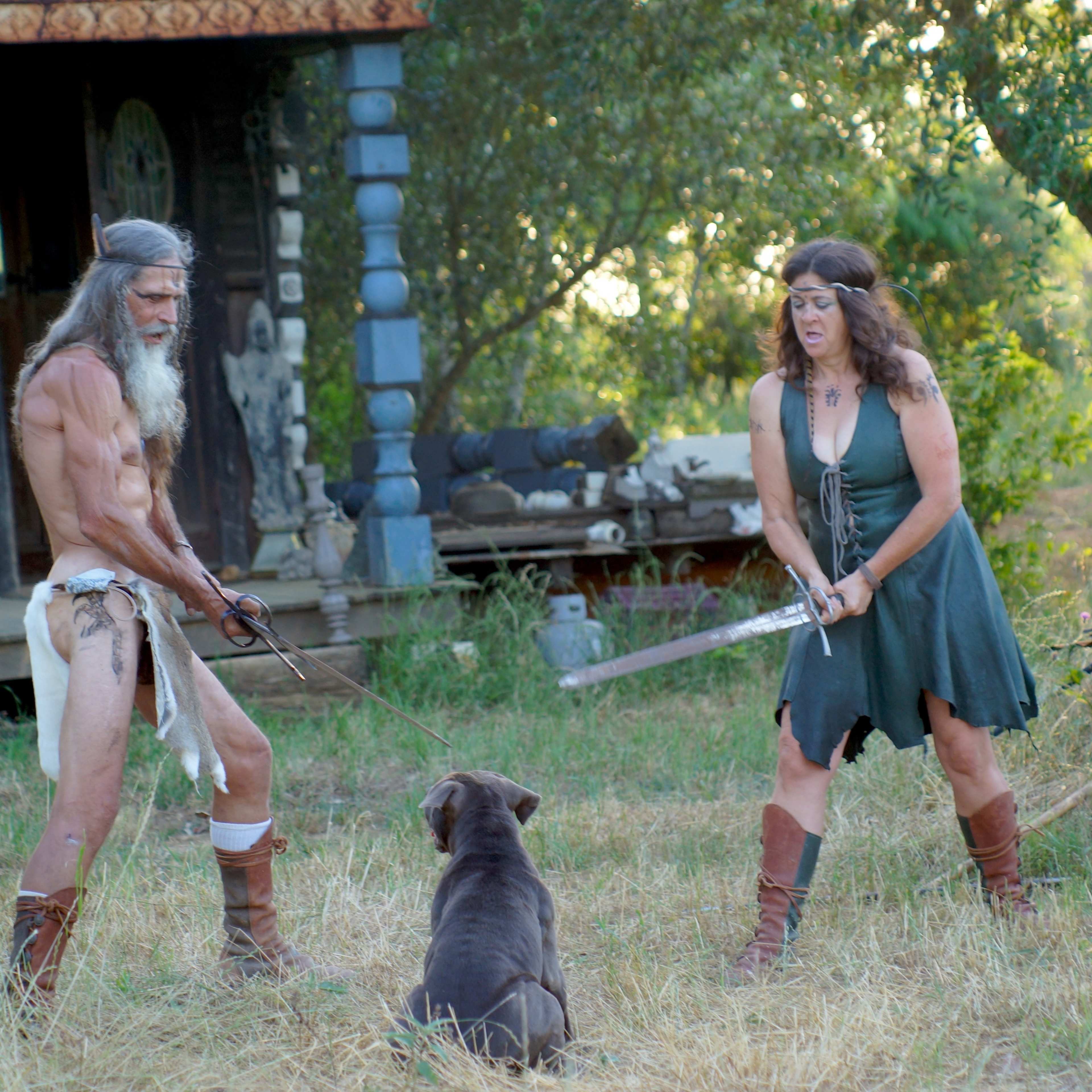 A man and a woman, both dressed in primitive attire, stand facing each other with swords while a dog sits between them in a grassy area near a rustic cabin.