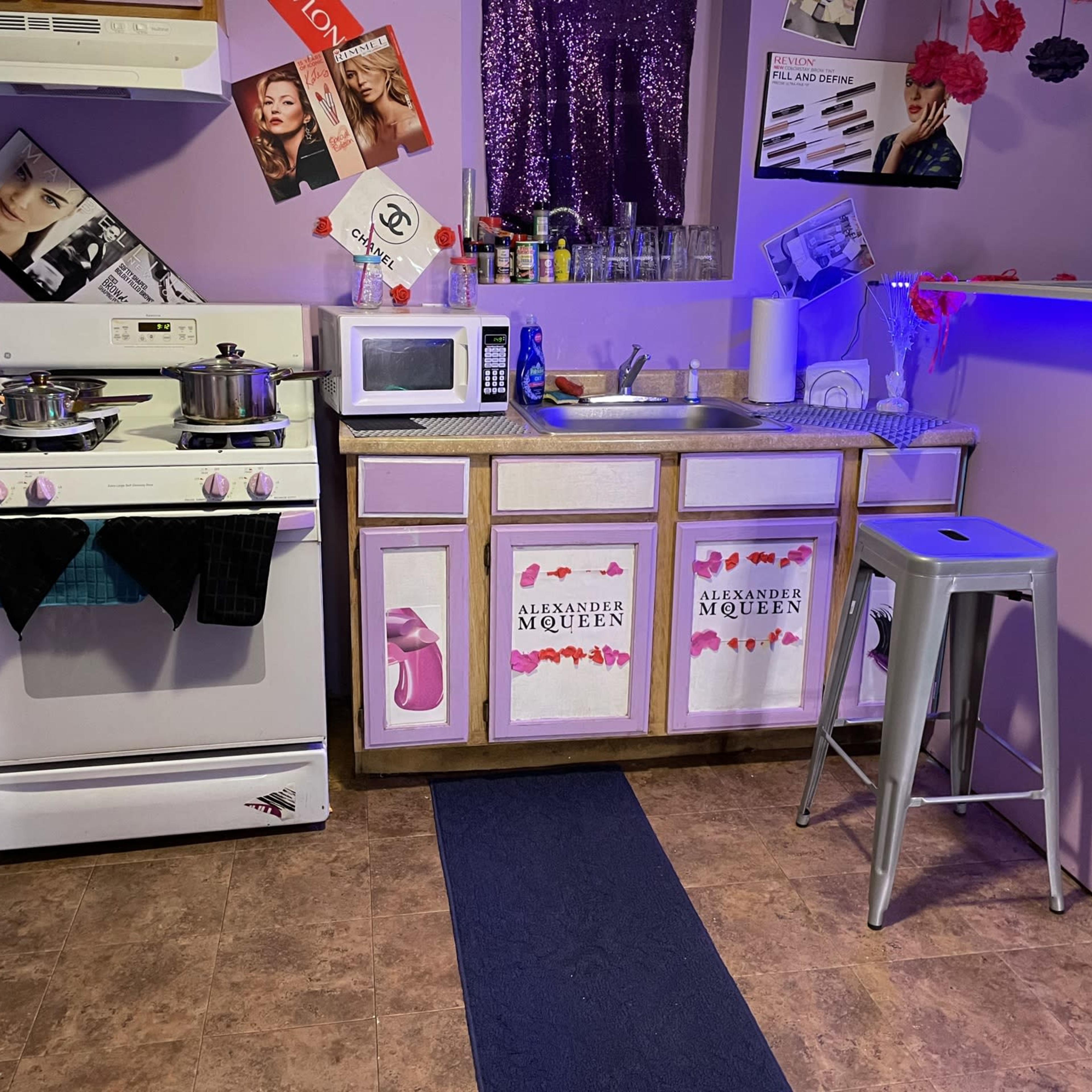 A small kitchen with a white stove, microwave, and a counter decorated with pink cabinets and various posters on the wall.