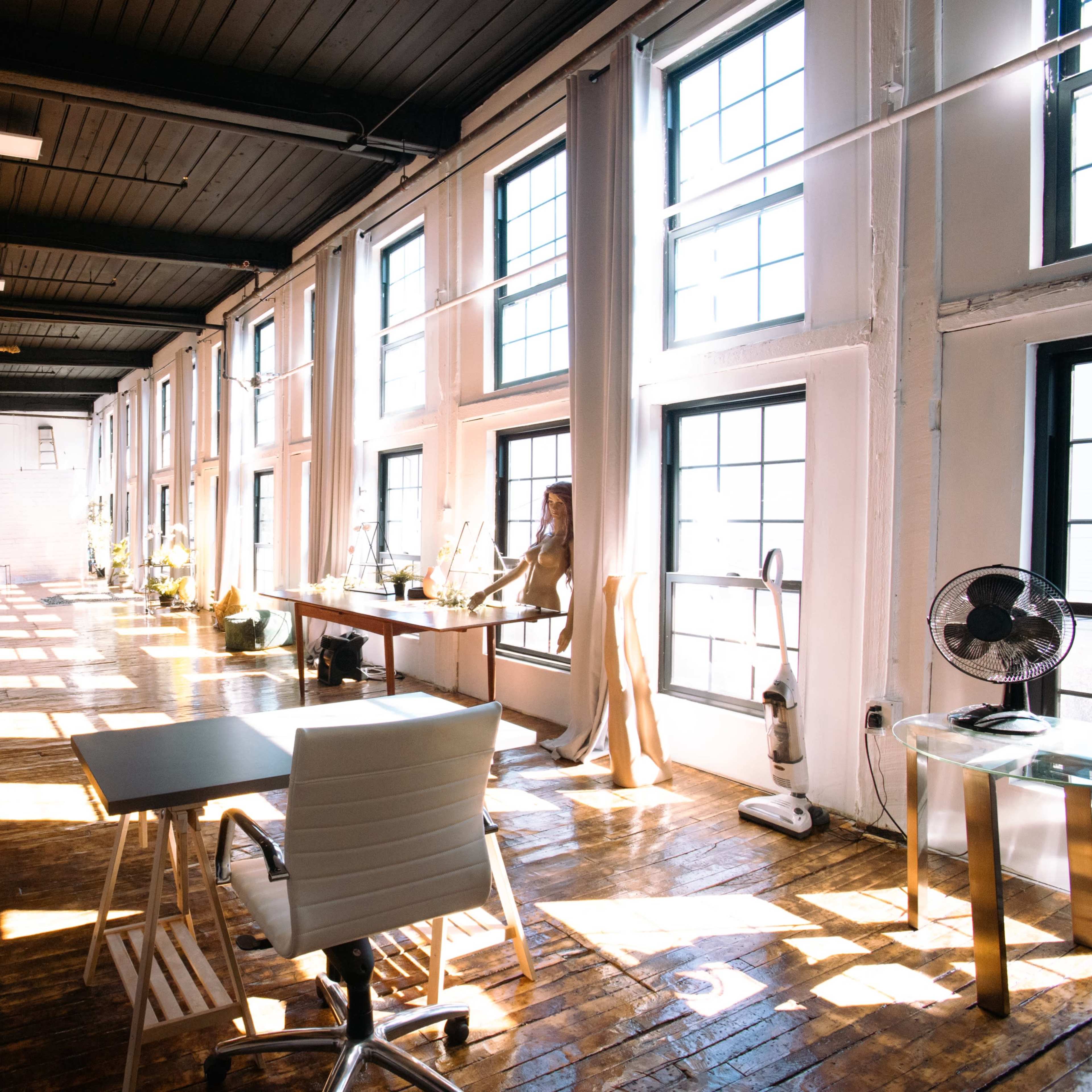 A spacious, brightly lit office with large windows, wooden floors, and minimal furniture, including a desk and an office chair.