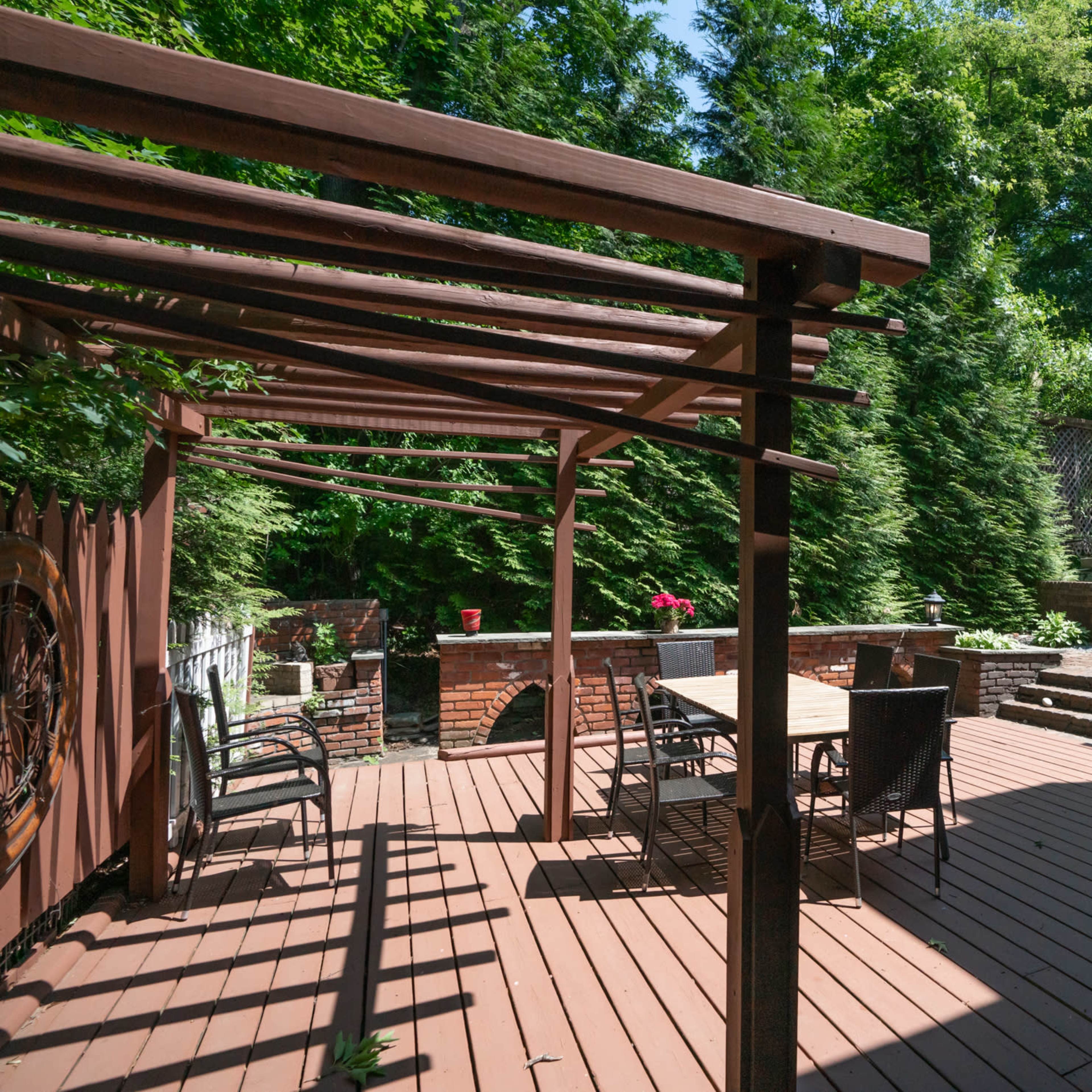 A wooden deck area with a pergola, dining table, and chairs surrounded by trees and garden landscaping.
