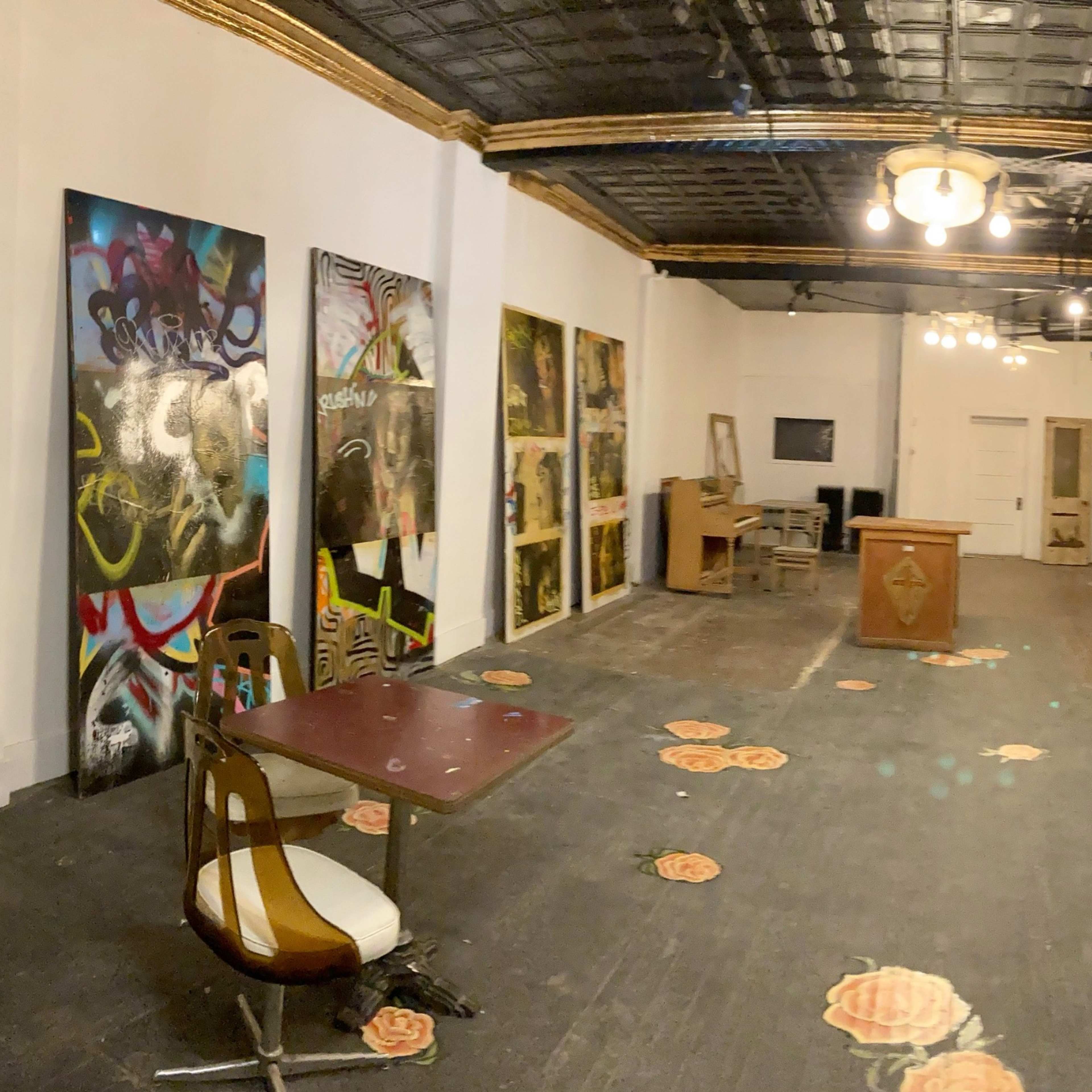 A spacious, well-lit room with a sofa, a table and chair, several large paintings on the wall, and floral-patterned flooring.