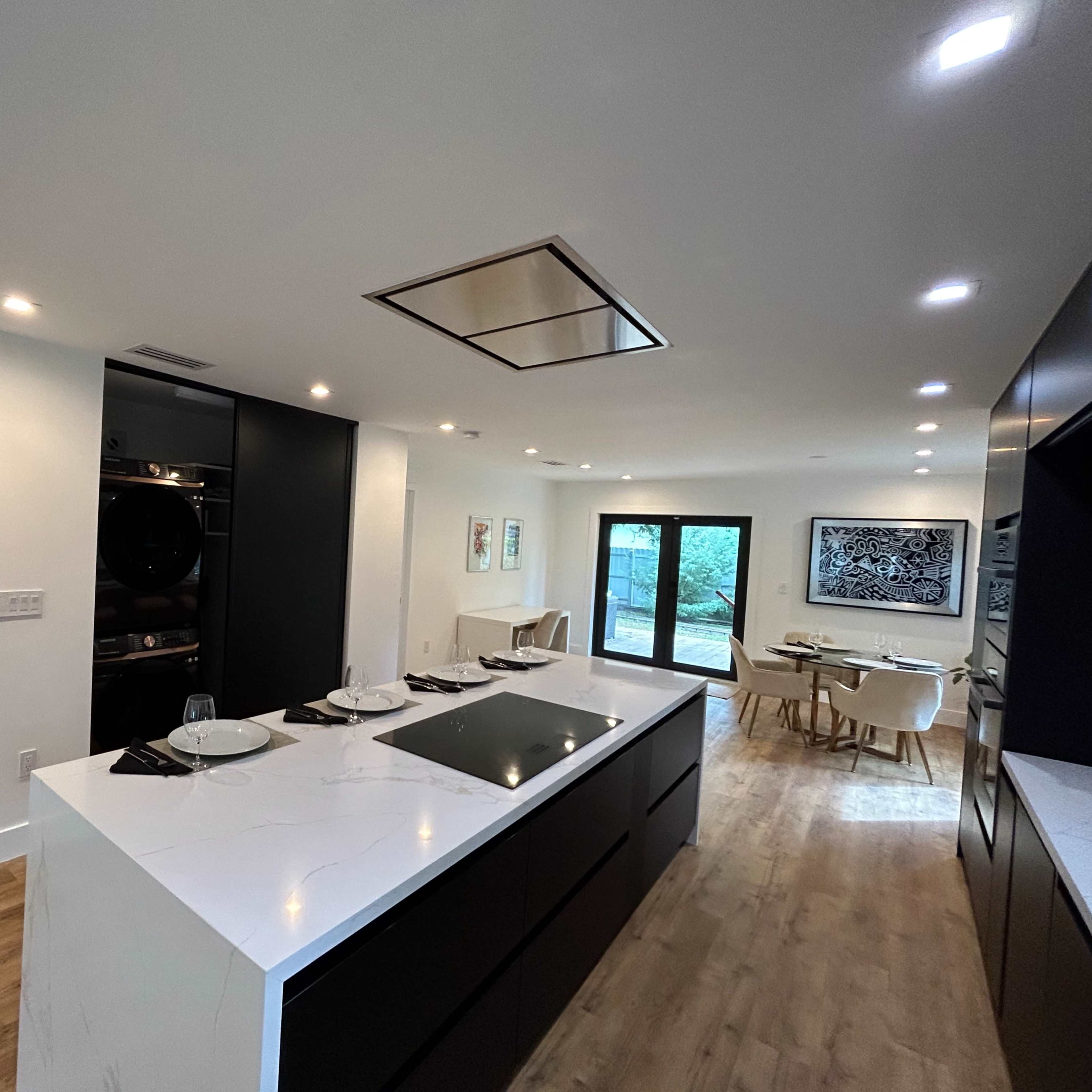 A modern kitchen featuring a large white island with a stovetop, black cabinetry, and a dining area with a table and chairs in the background.