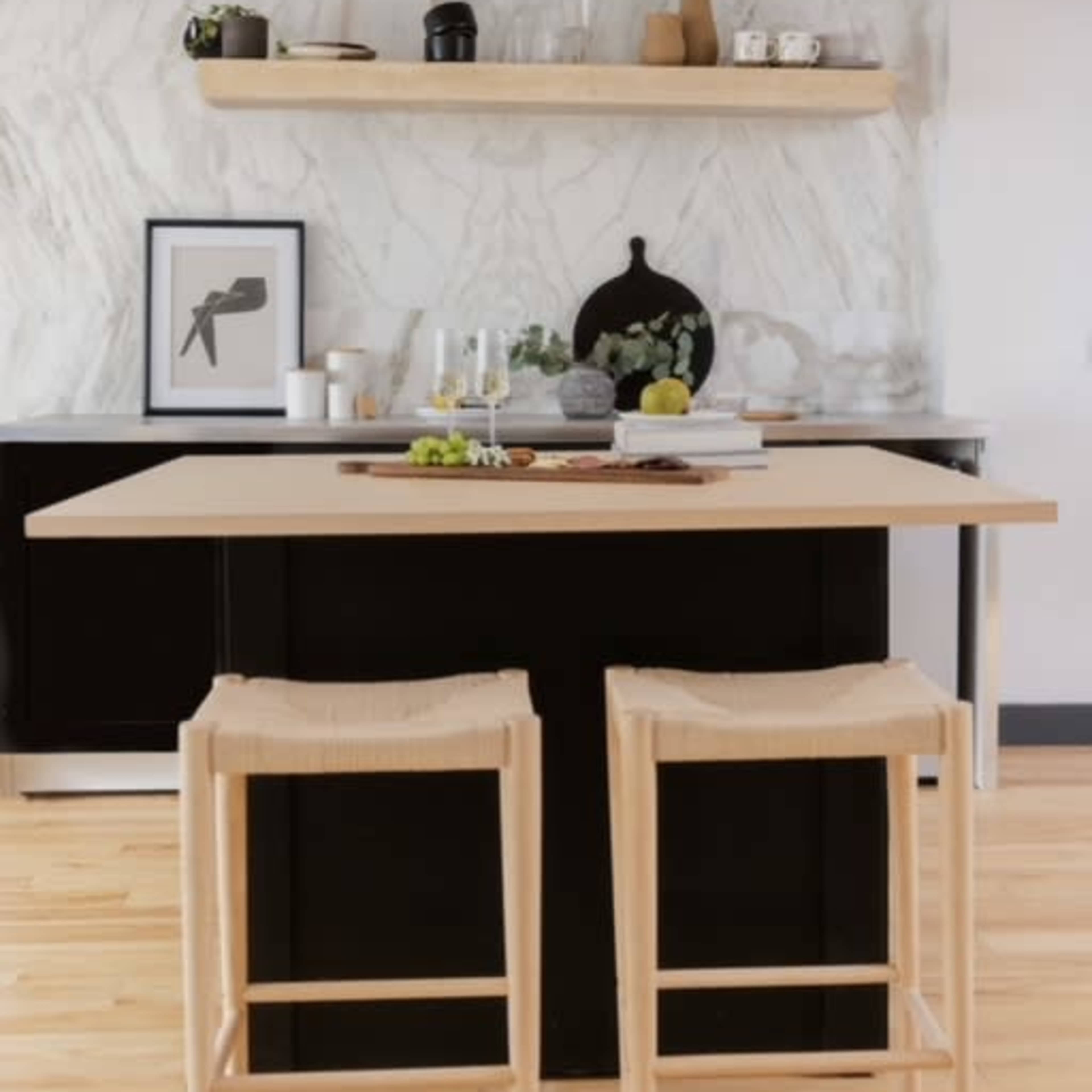 A modern kitchen with a black island, two light-colored stools, a marble backsplash, and a shelf displaying kitchen items.