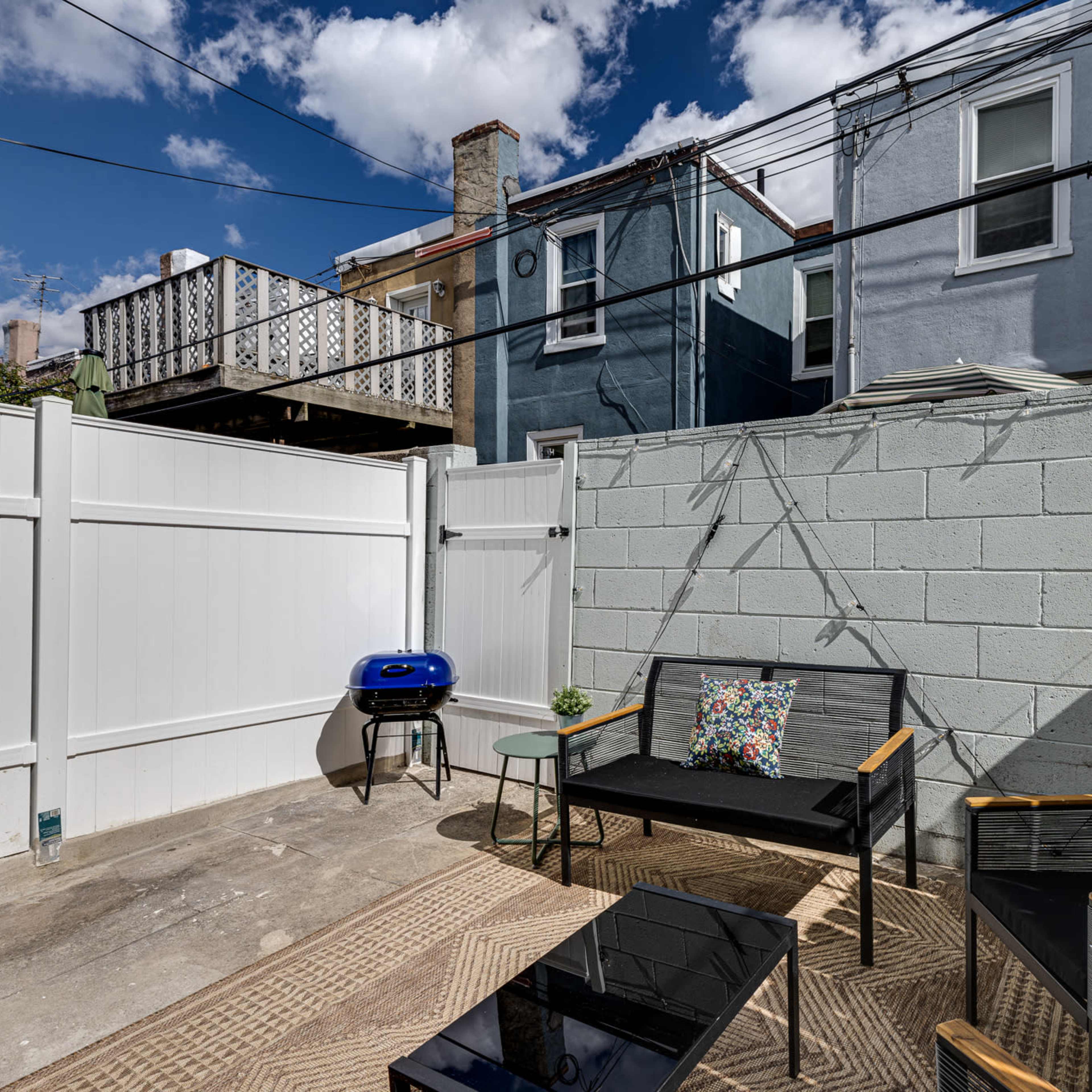 A small, enclosed patio features a black table with chairs, a blue barbecue grill, and a concrete wall surrounded by buildings and power lines under a partly cloudy sky.