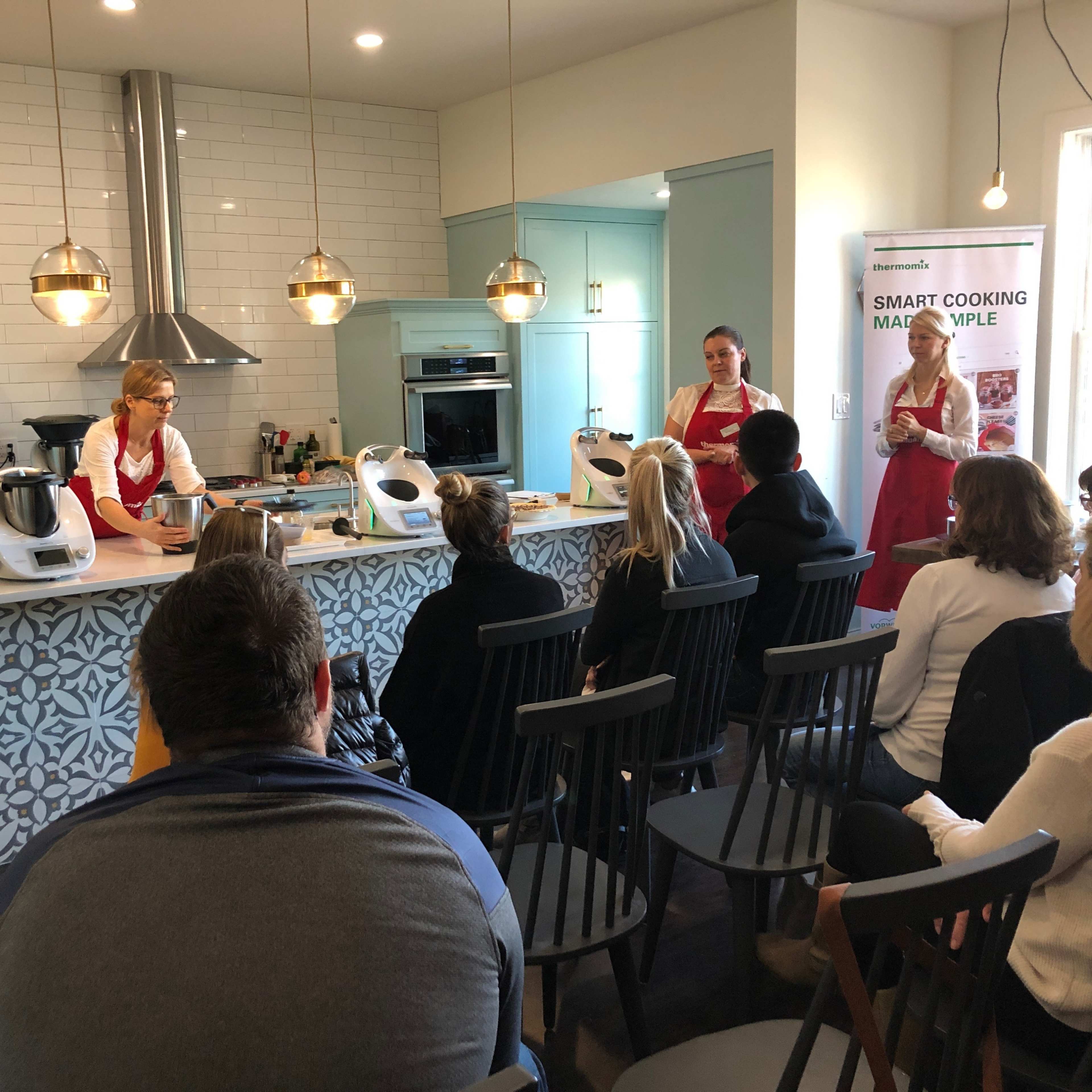 A group of people sits on chairs facing a kitchen where two instructors present a cooking demonstration.