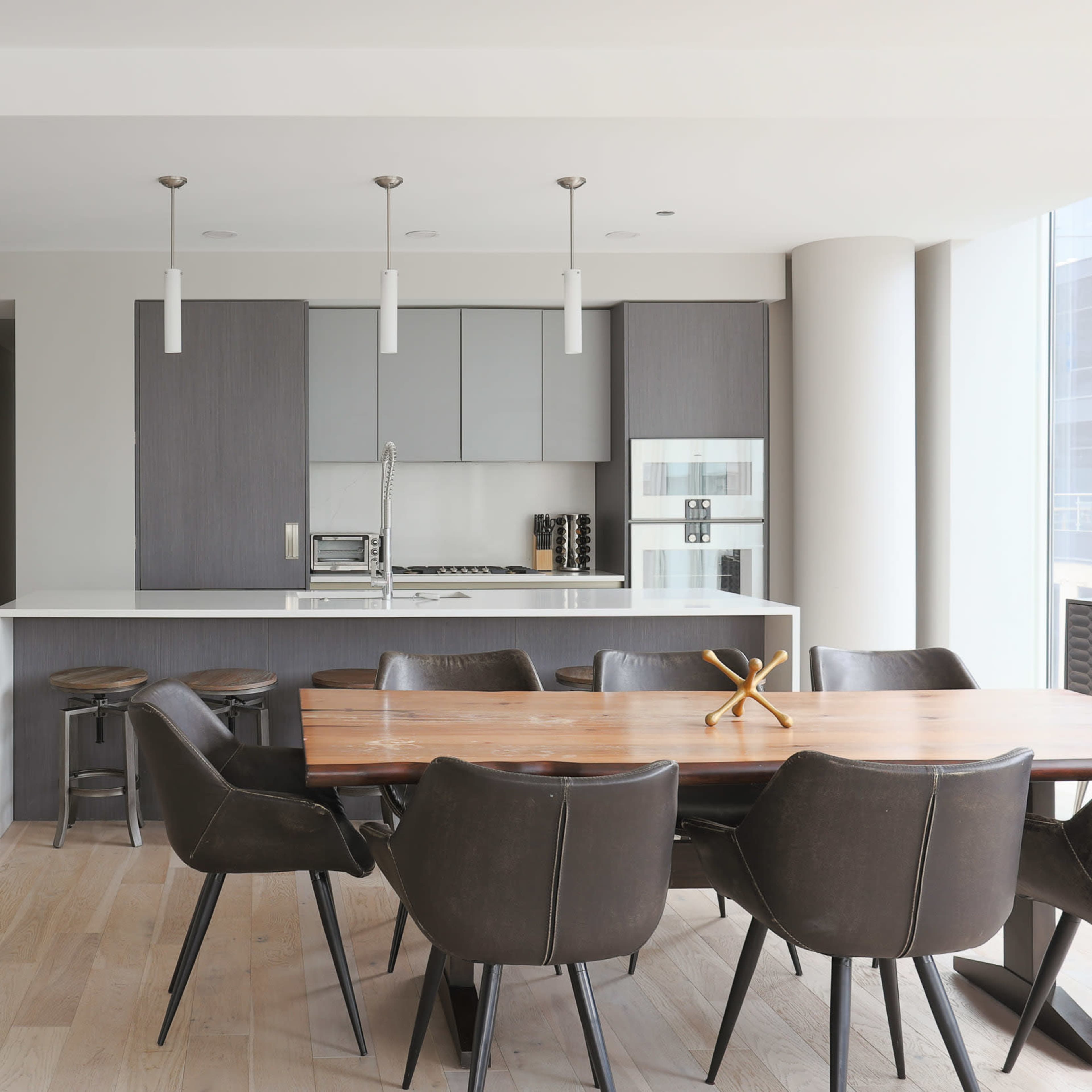 A modern kitchen and dining area featuring a large wooden table surrounded by black chairs, with minimalist cabinetry and pendant lighting above.