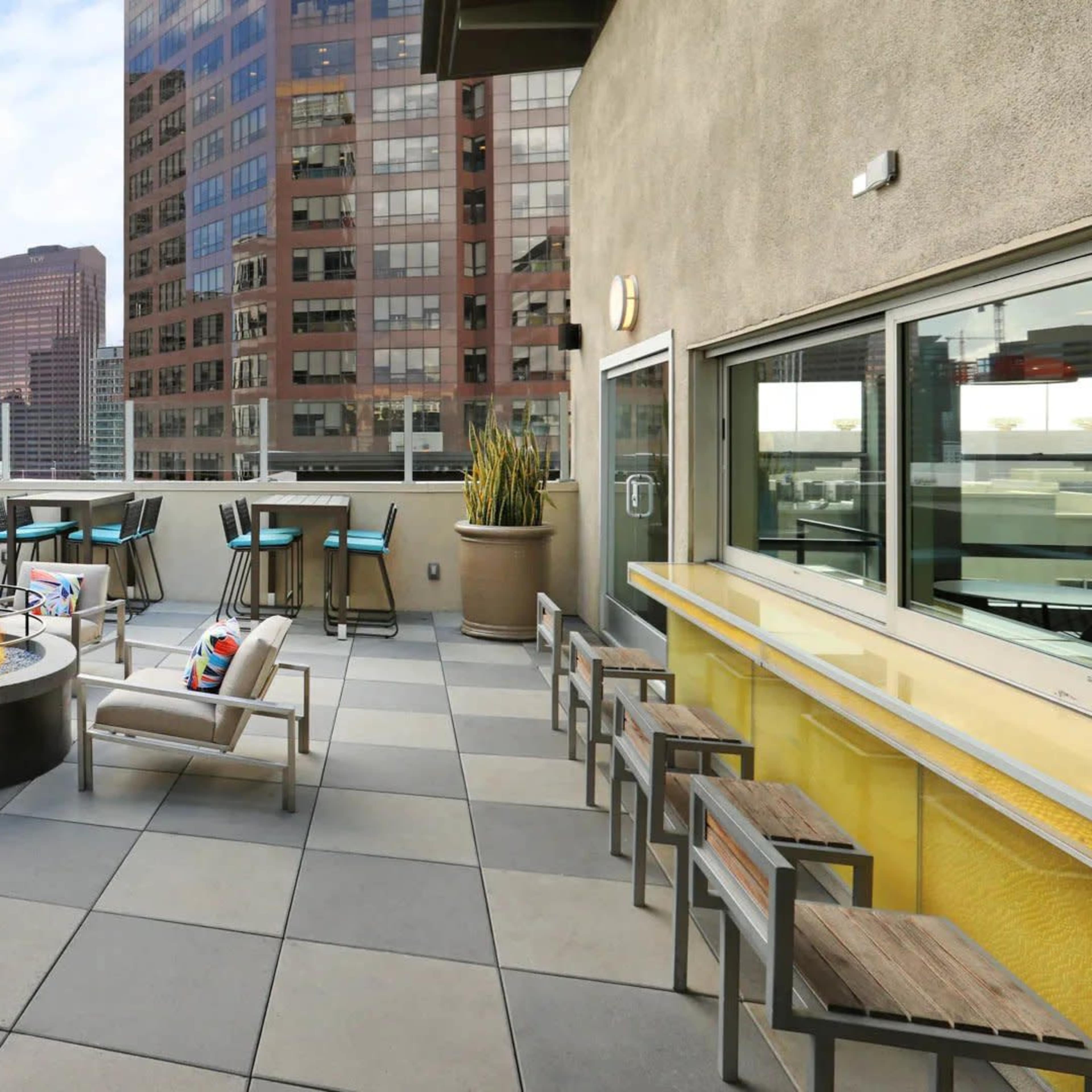 A rooftop terrace featuring modern seating around a circular fire pit, with tall buildings and a clear sky in the background.