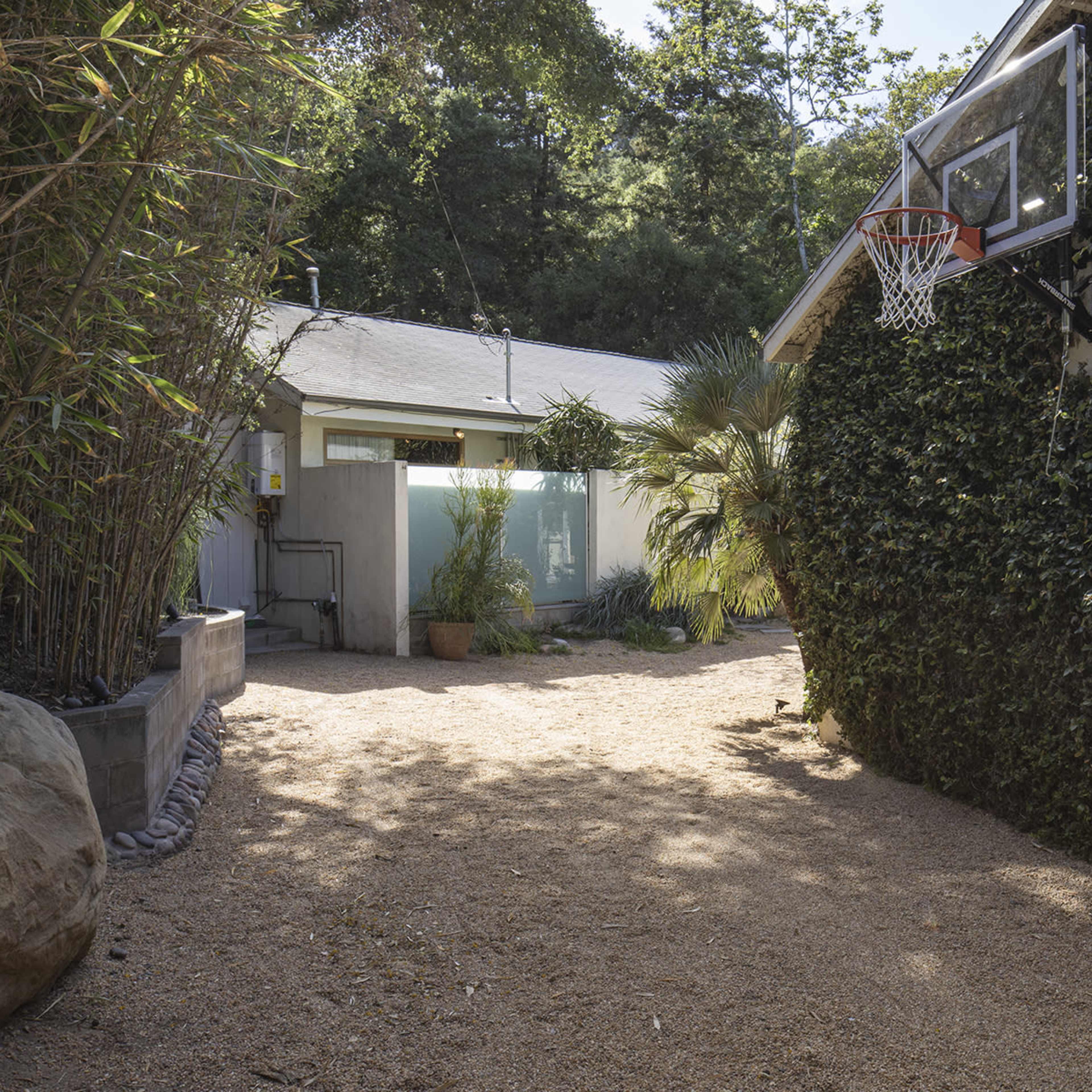 The scene features a gravel pathway leading to a house on one side and a basketball hoop attached to a wall on the other, surrounded by greenery and bamboo.