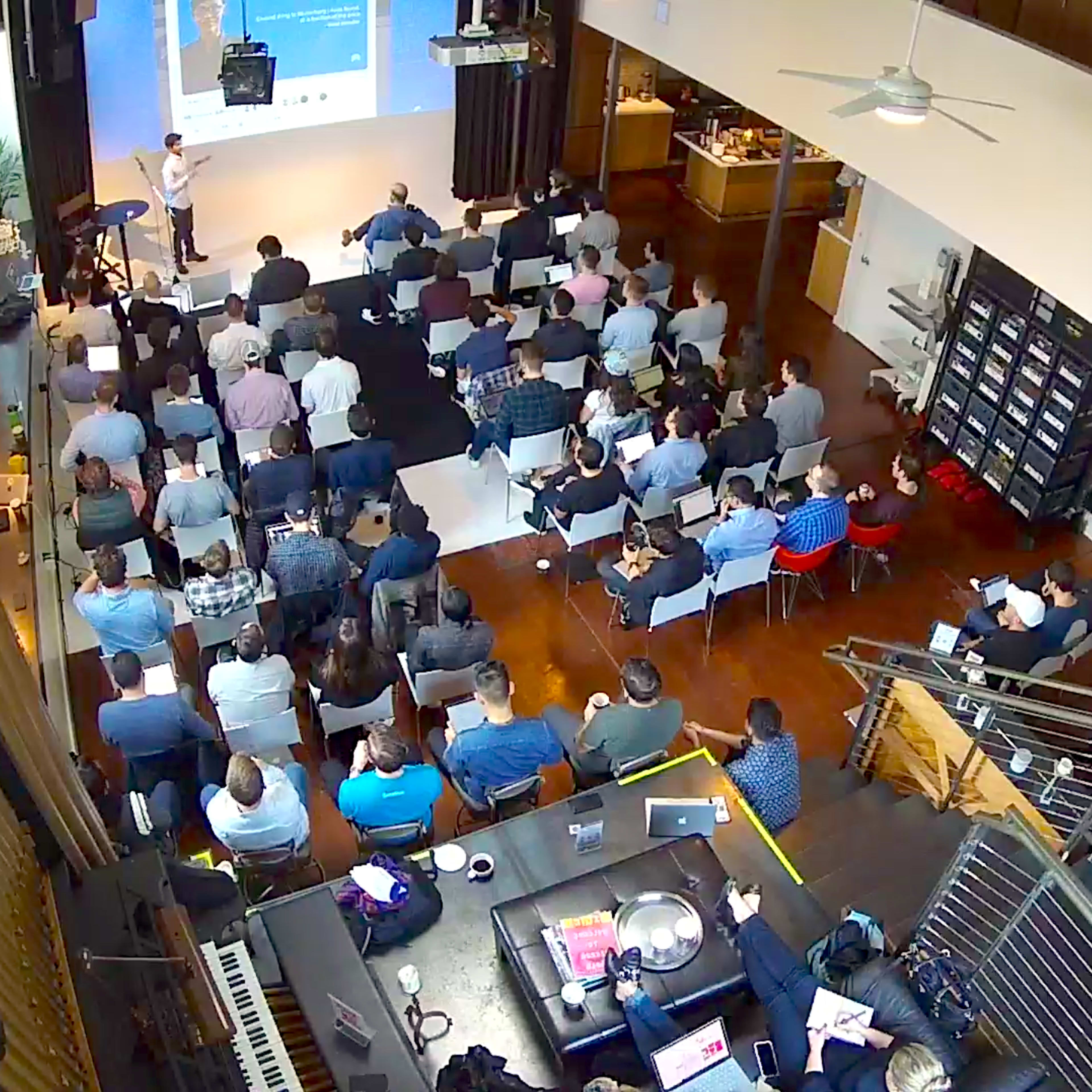 A speaker presents to an audience seated in a spacious room with large windows and wooden flooring.