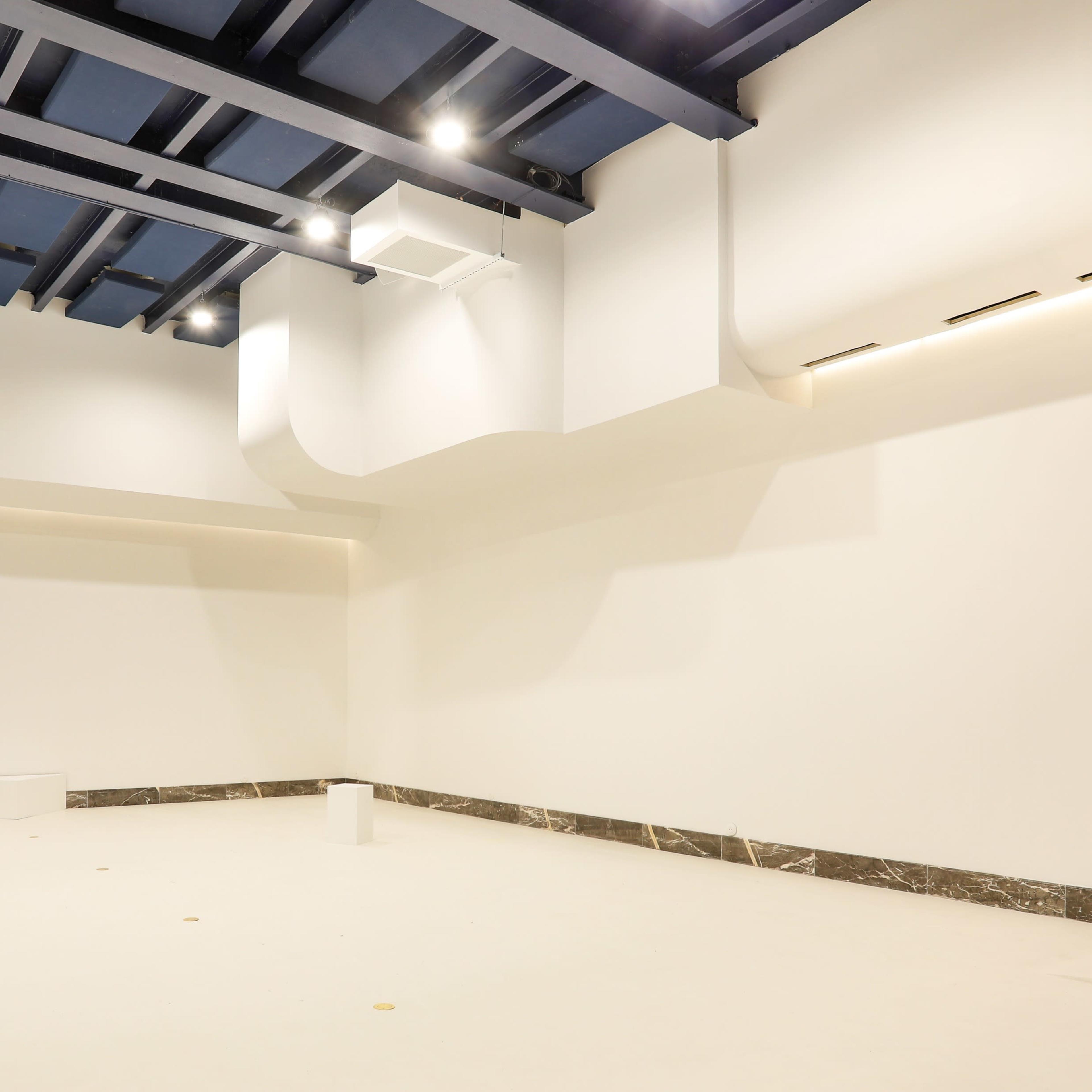 A minimalist interior space with white walls, a light-colored floor, and blue ceiling beams, featuring a few white cylindrical forms scattered throughout.