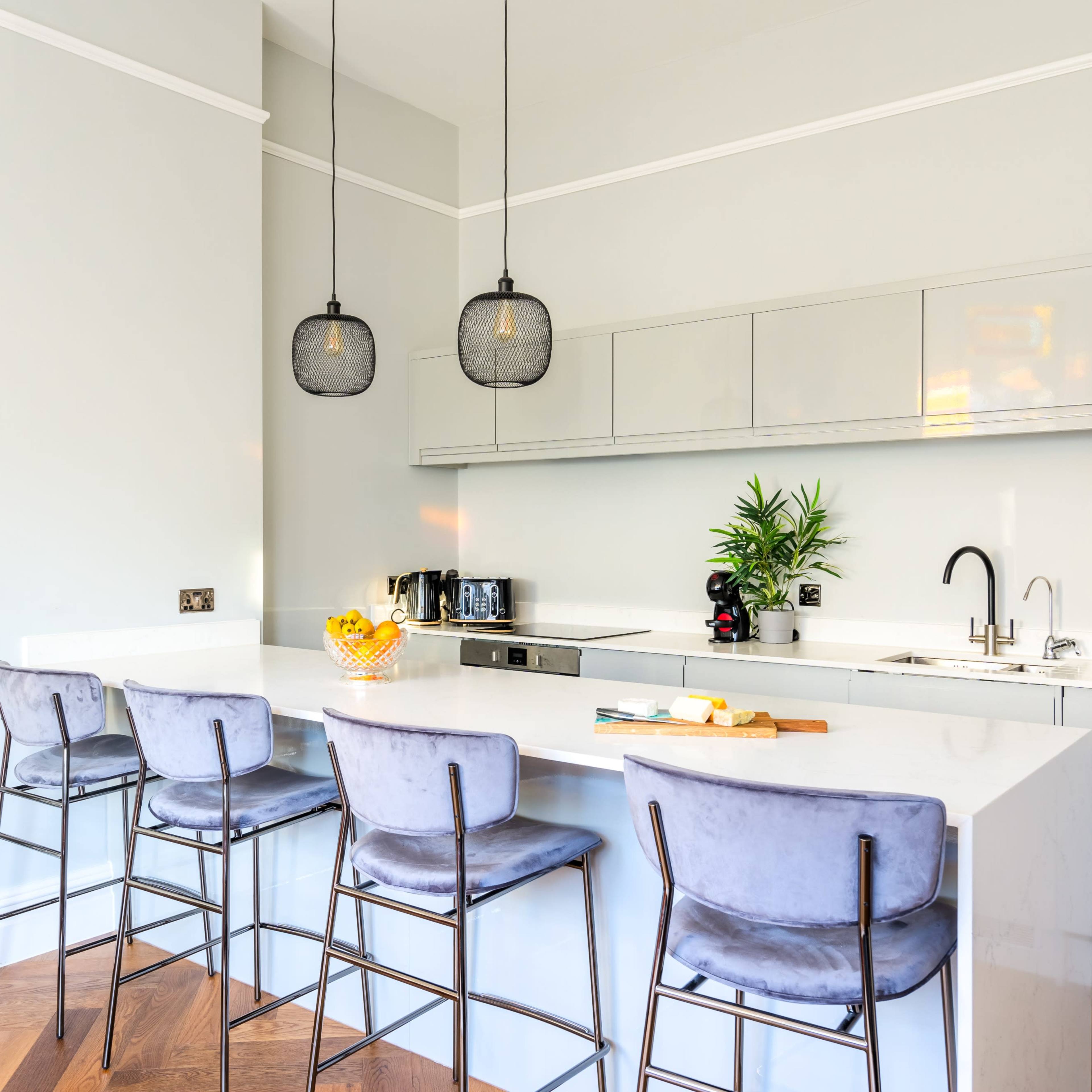 A modern kitchen features a white countertop with four high-backed stools, pendant lights overhead, and minimalist cabinetry along the wall.