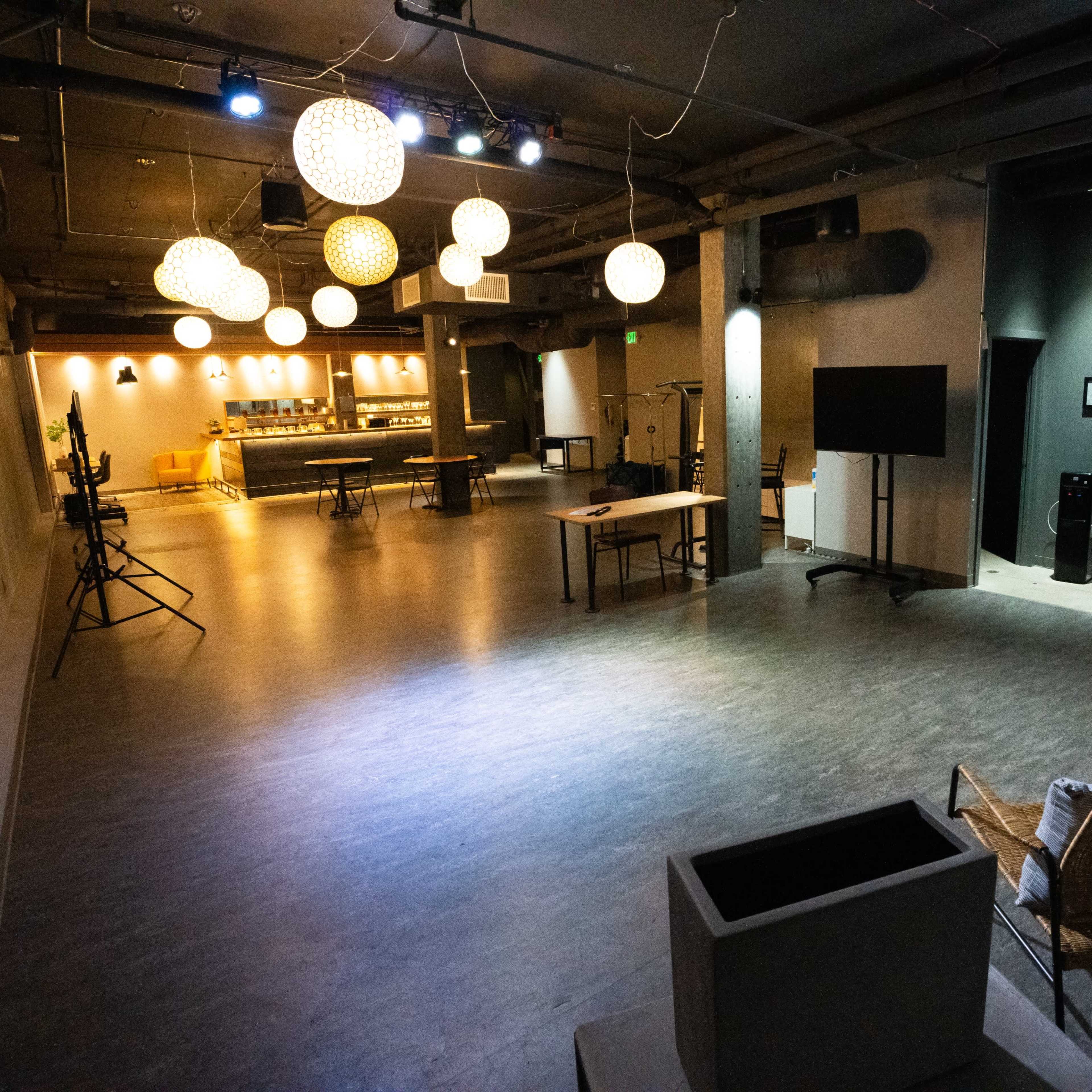 A spacious, modern indoor venue with light fixtures hanging from the ceiling, a bar area in the background, and a few seating arrangements in the foreground.