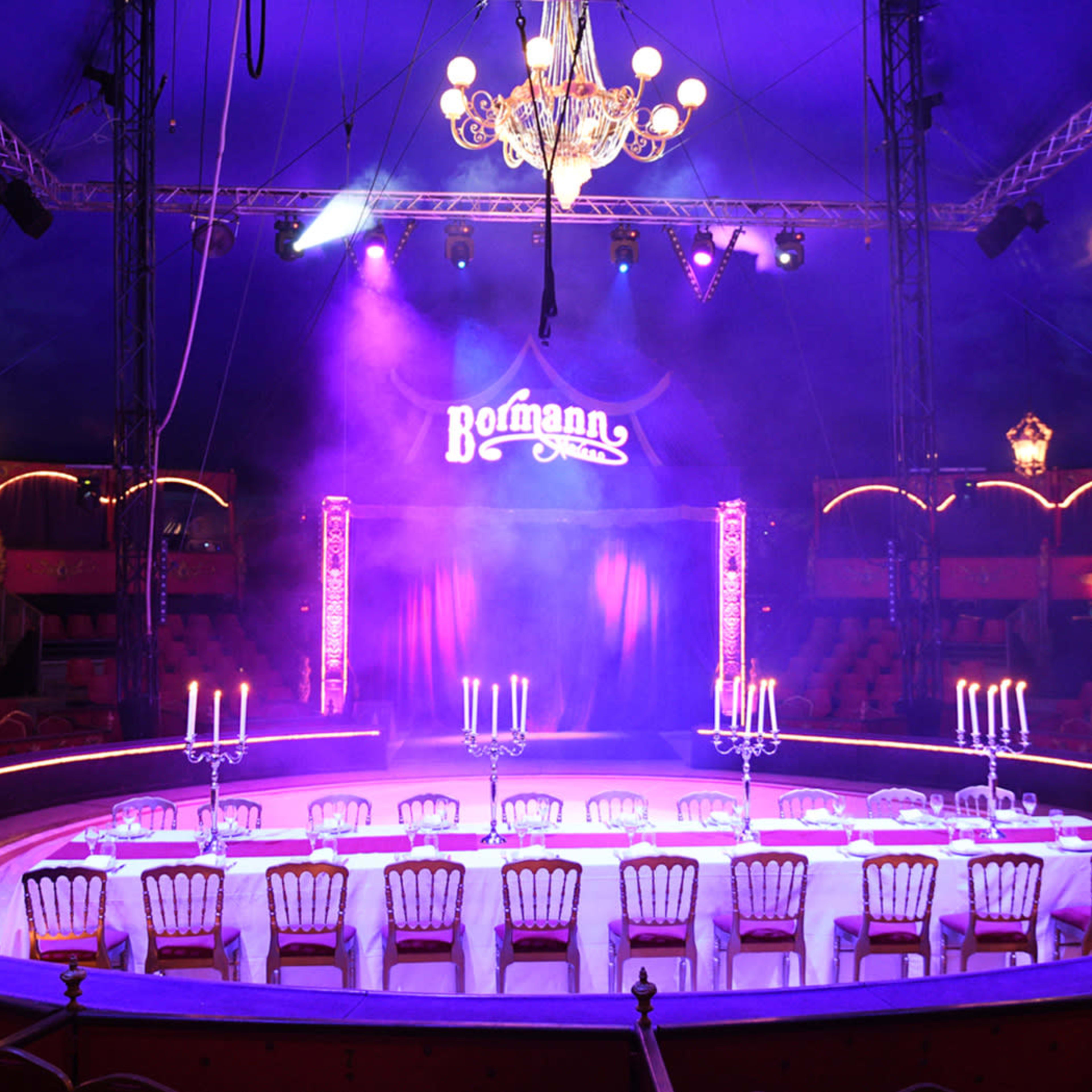 A brightly lit circular stage set up with a long table and chairs, surrounded by empty seats in a circus tent.