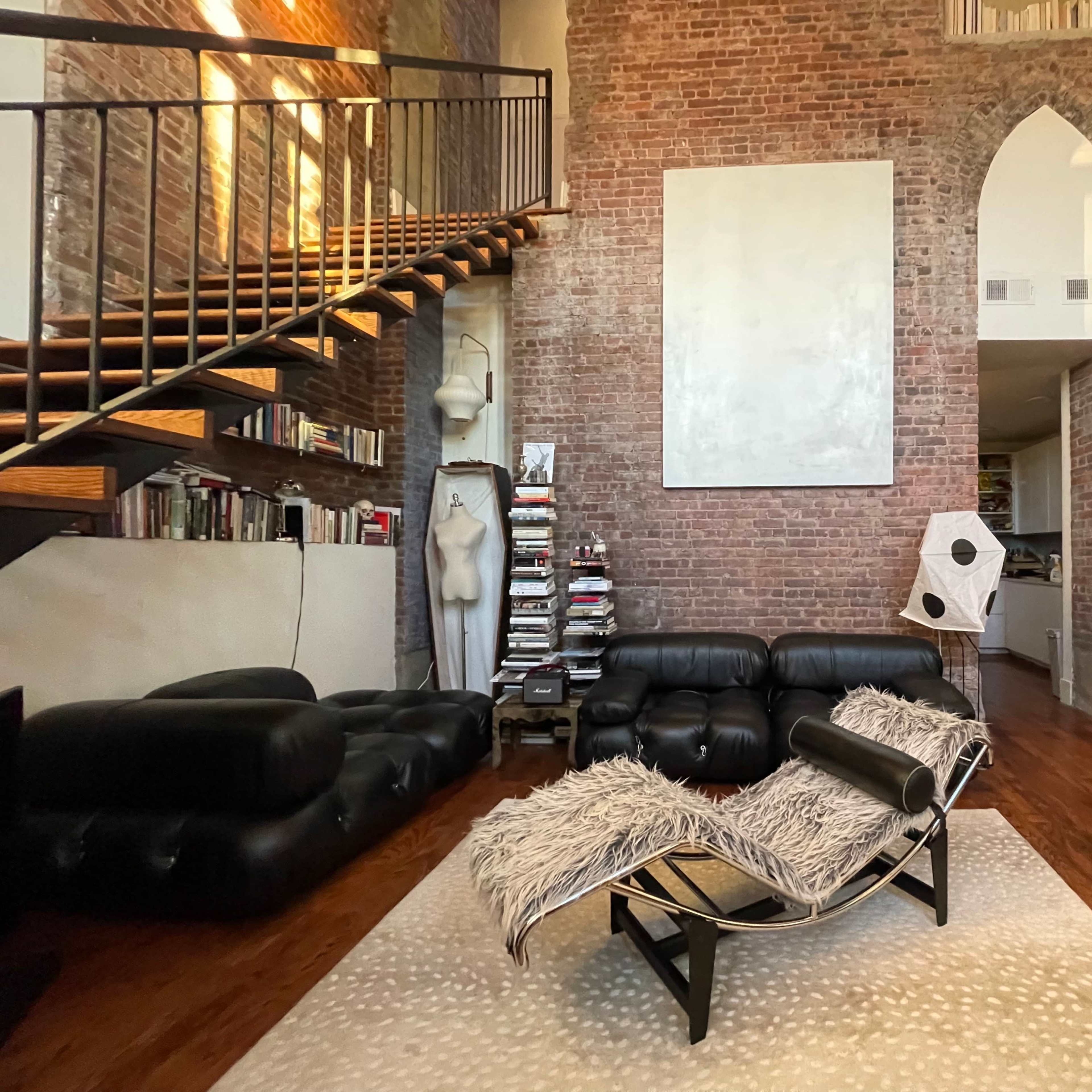 The room features a modern lounge area with a black leather sofa, a unique lounge chair, and a staircase leading to a loft, all set against exposed brick walls.