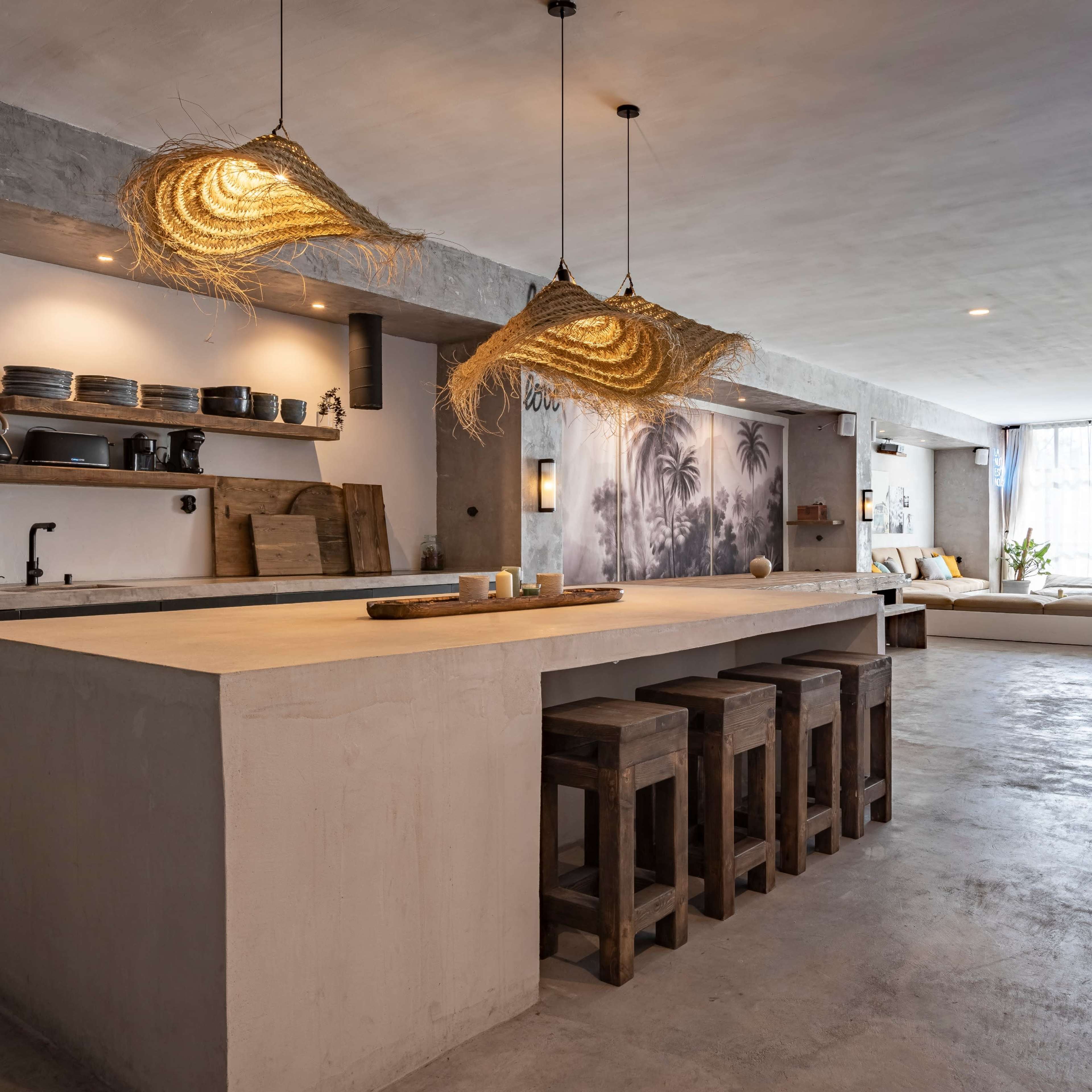 A modern kitchen and living area with a large concrete island, wooden stools, and hanging straw lights, leading to a seating area with a large window.