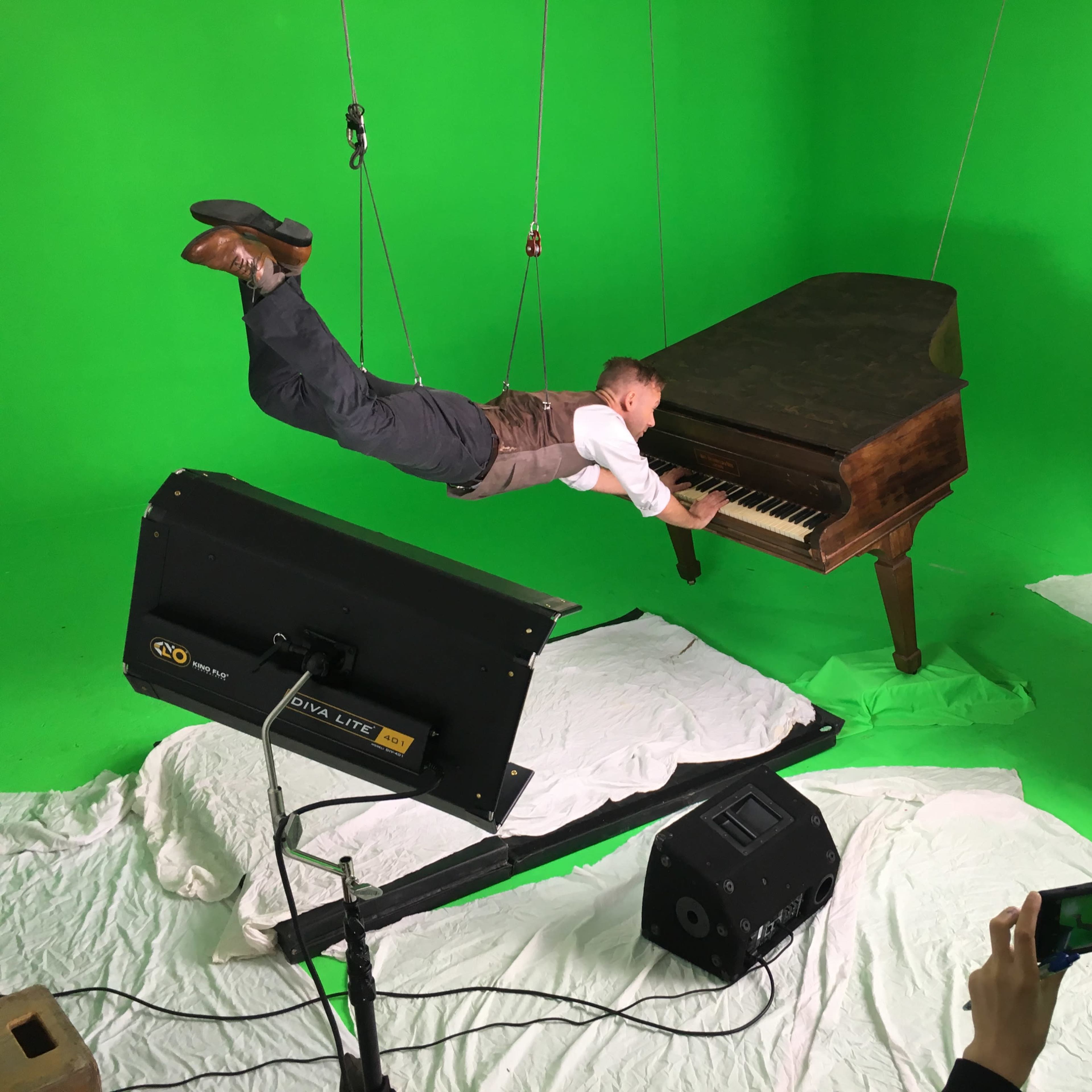 A person in a suit is suspended mid-air while playing a grand piano against a green screen backdrop.