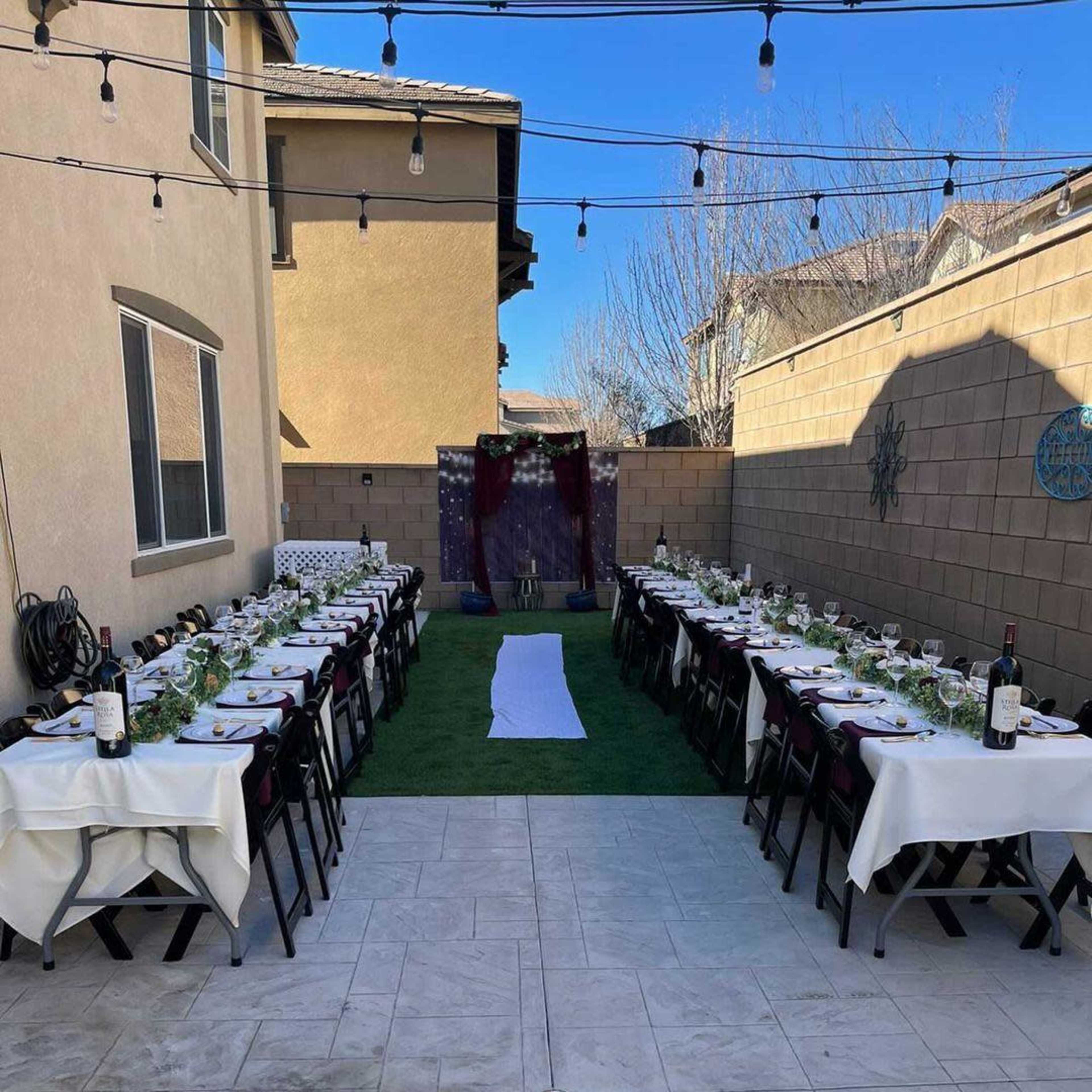 A long, elegantly set outdoor dining table is arranged on grass between two buildings, with string lights overhead and decorative elements in the background.