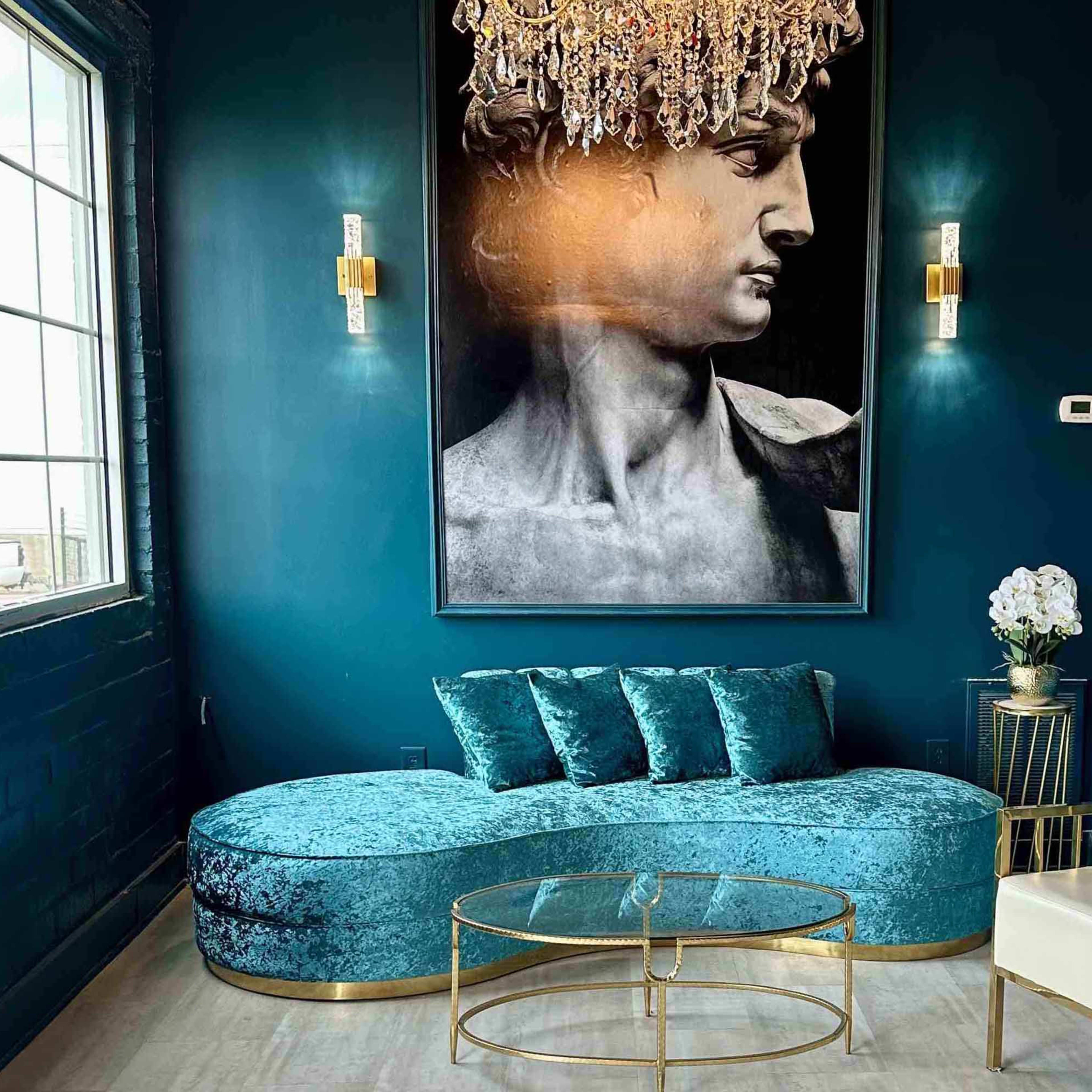 A modern lounge area featuring a teal curved sofa, a glass coffee table, a gold-framed chair, and a large black-and-white portrait of a male figure on the wall under a chandelier.