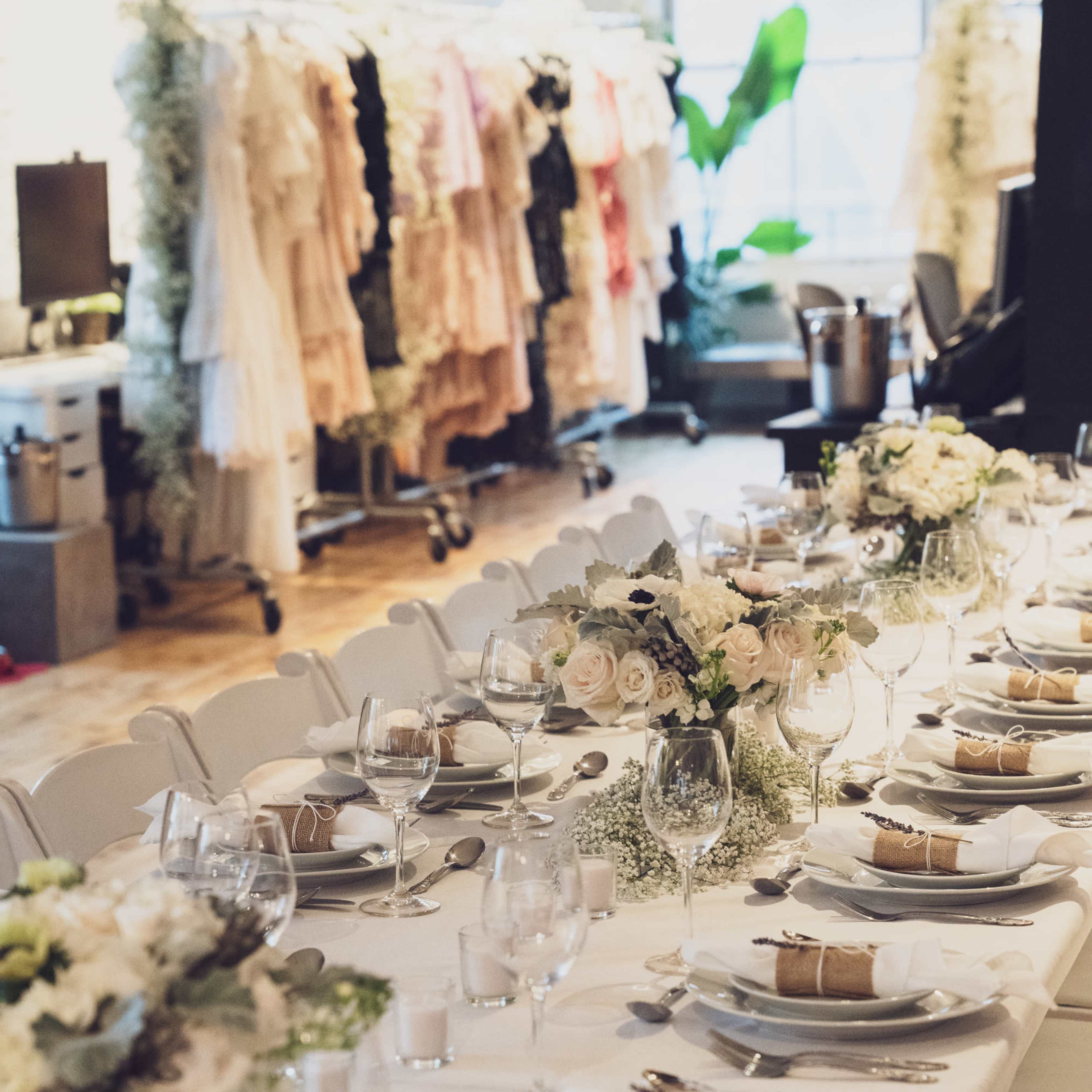 A long dining table is elegantly set with white plates, silverware, and floral centerpieces, while a collection of dresses hangs in the background.