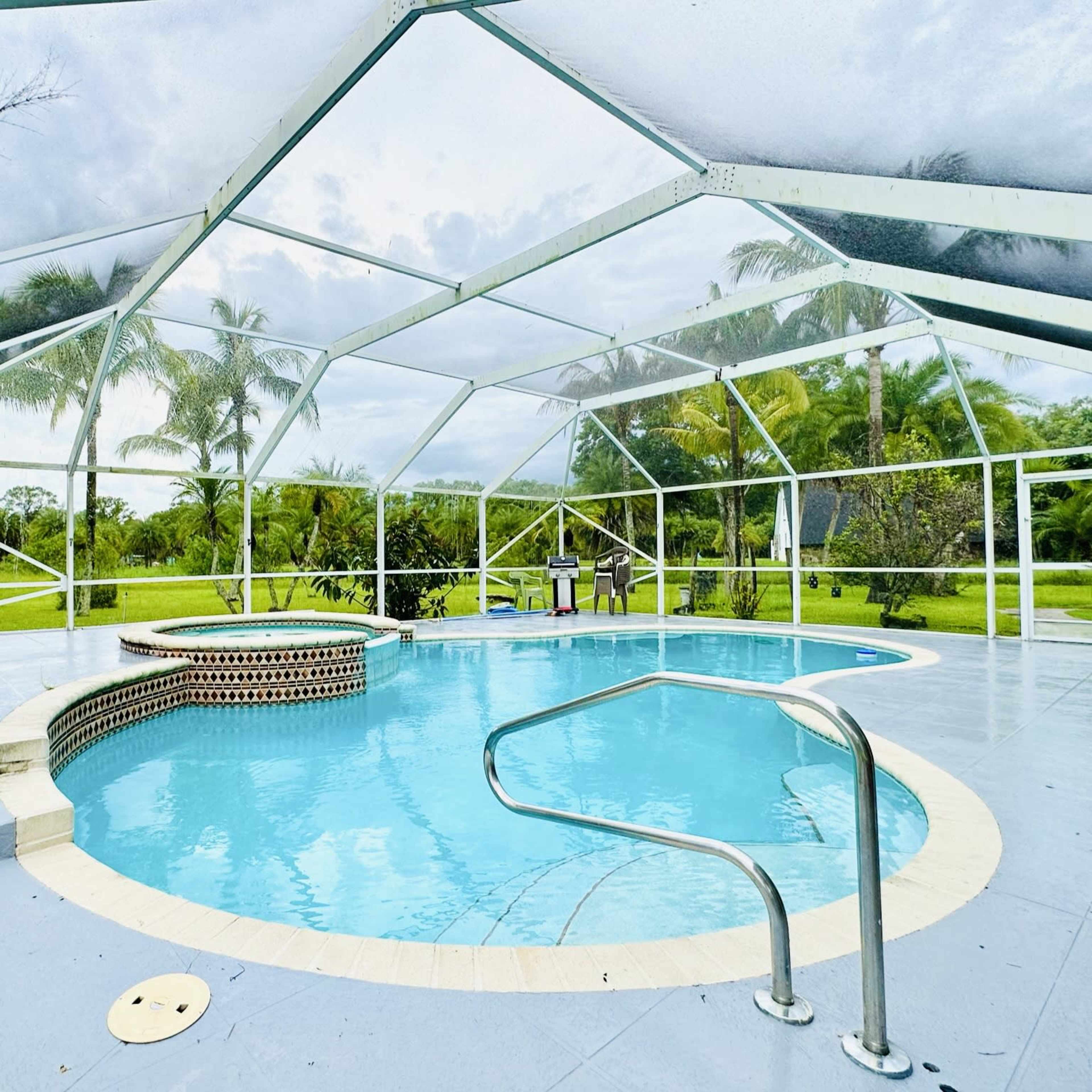 A screened-in outdoor pool area features a round hot tub, lounge chairs, and views of a green landscape.
