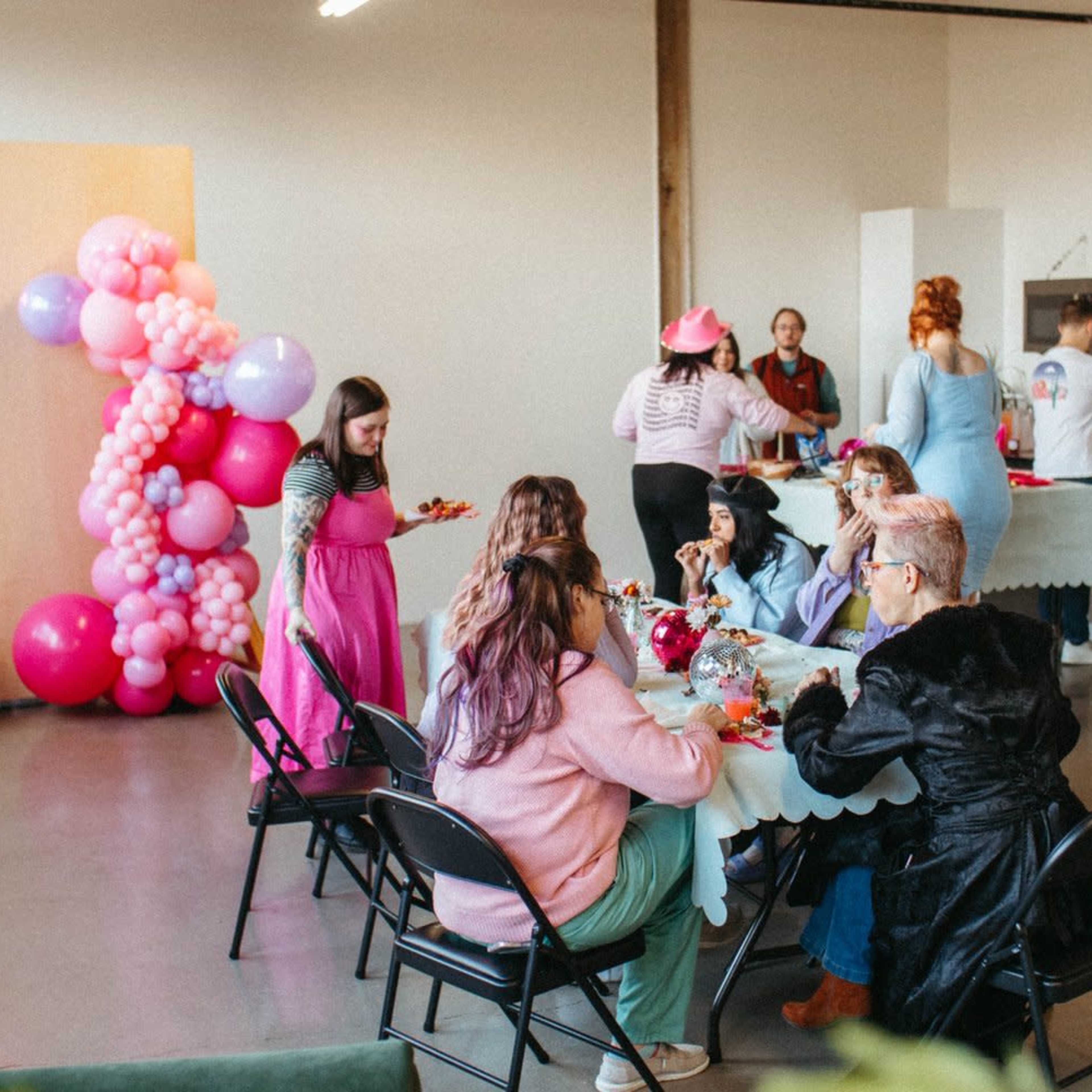 A group of people sits at a table enjoying food in a well-lit, spacious room decorated with colorful balloons.