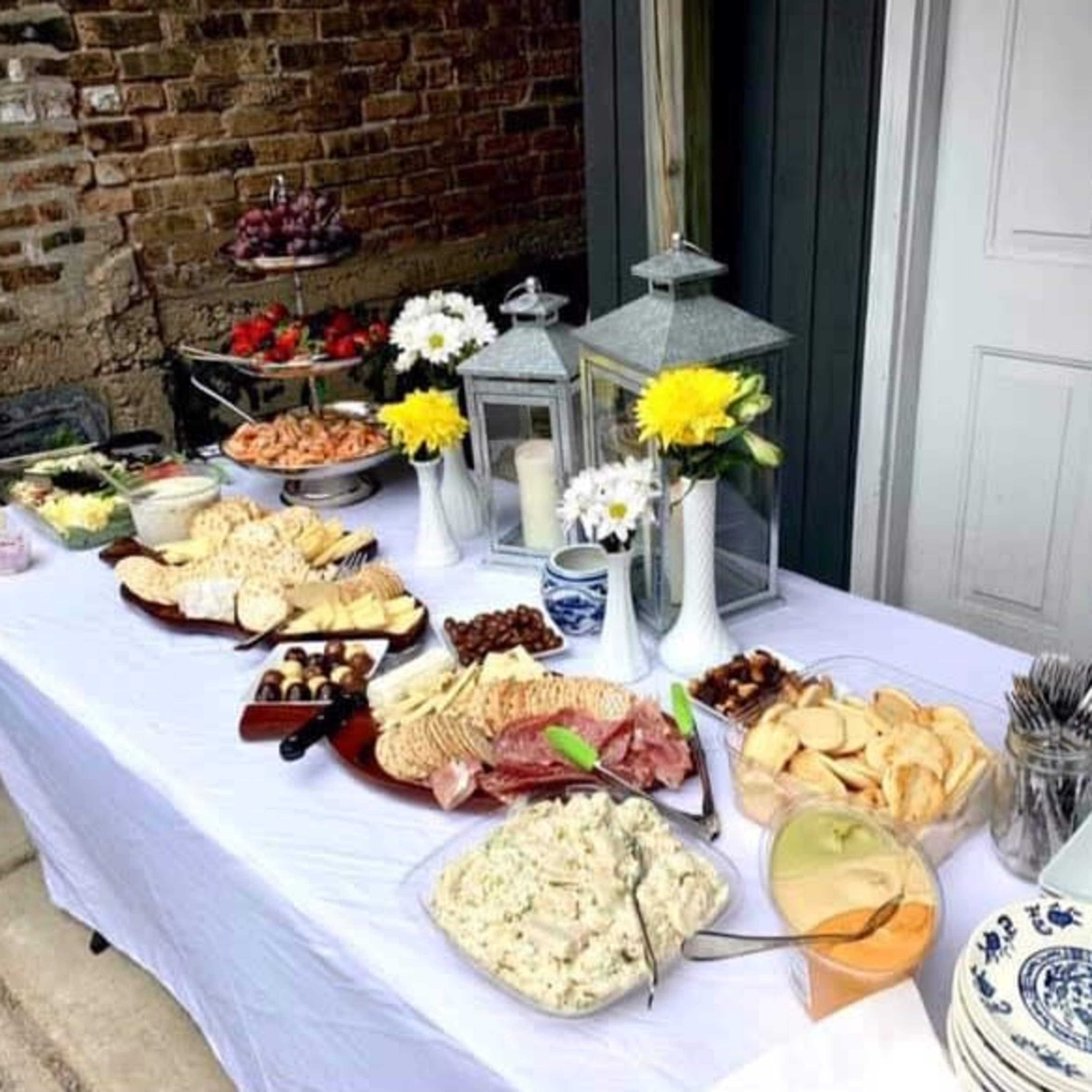 A food buffet is set up on a table featuring various platters of meats, cheeses, vegetables, and salads, along with drinks and decorative centerpieces.